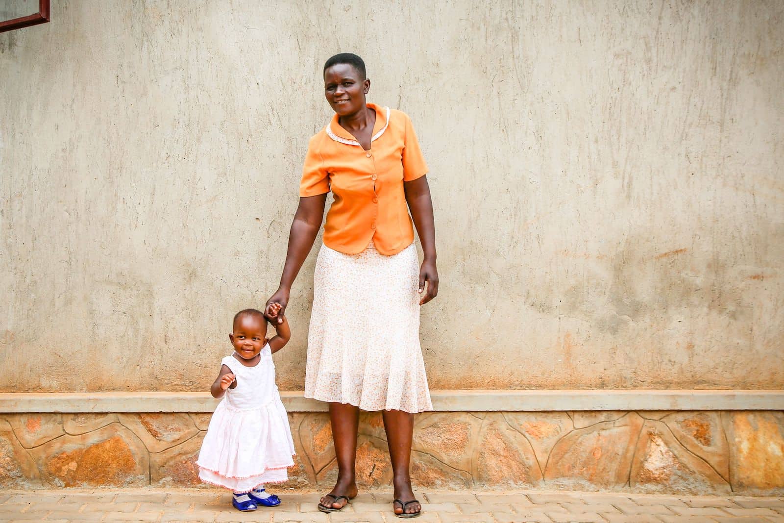 A woman in an orange shirt and white skirt stands in front of a beige wall, holding the hand of a baby in a white dress standing next to her.