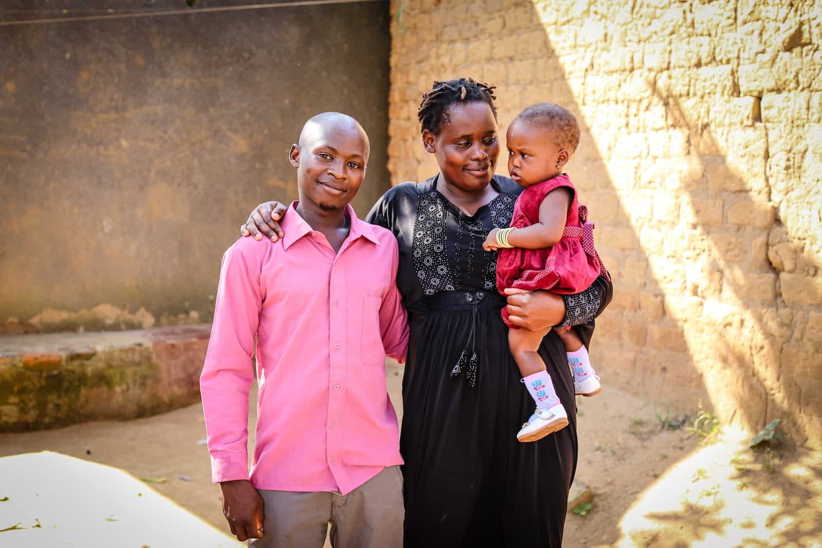 A man in a pink shirt with his arm around a woman in a black dress holding a baby in a red dress.