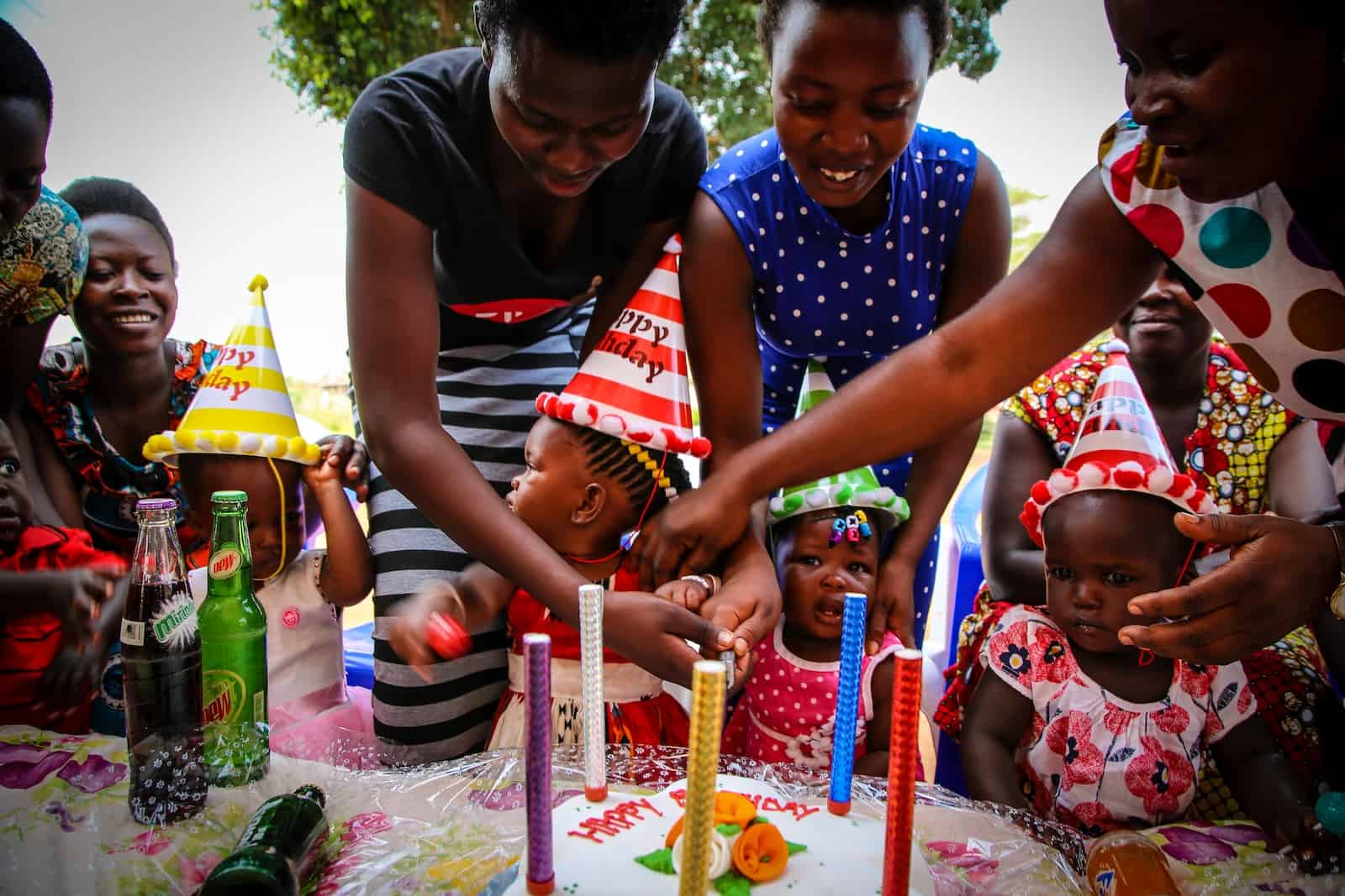 Three babies sit at a table with a cake in front of them to celebrate a birthday milestone. They are wearing party hats and women stand behind them, helping them.