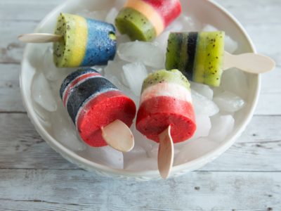 A white bowl full of ice holds five multi-colored popsicles, on a gray table.