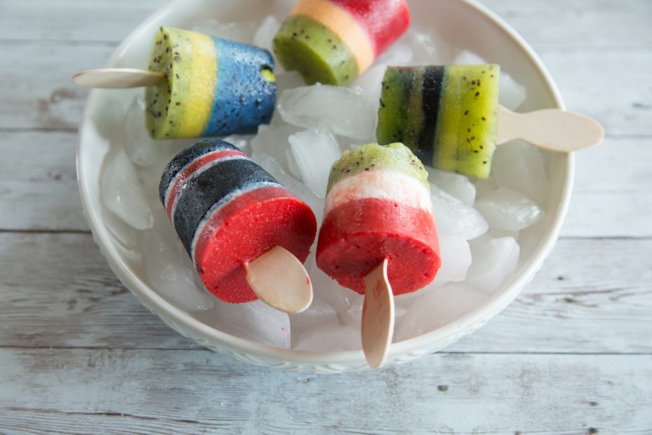 A white bowl full of ice holds five multi-colored popsicles, on a gray table.
