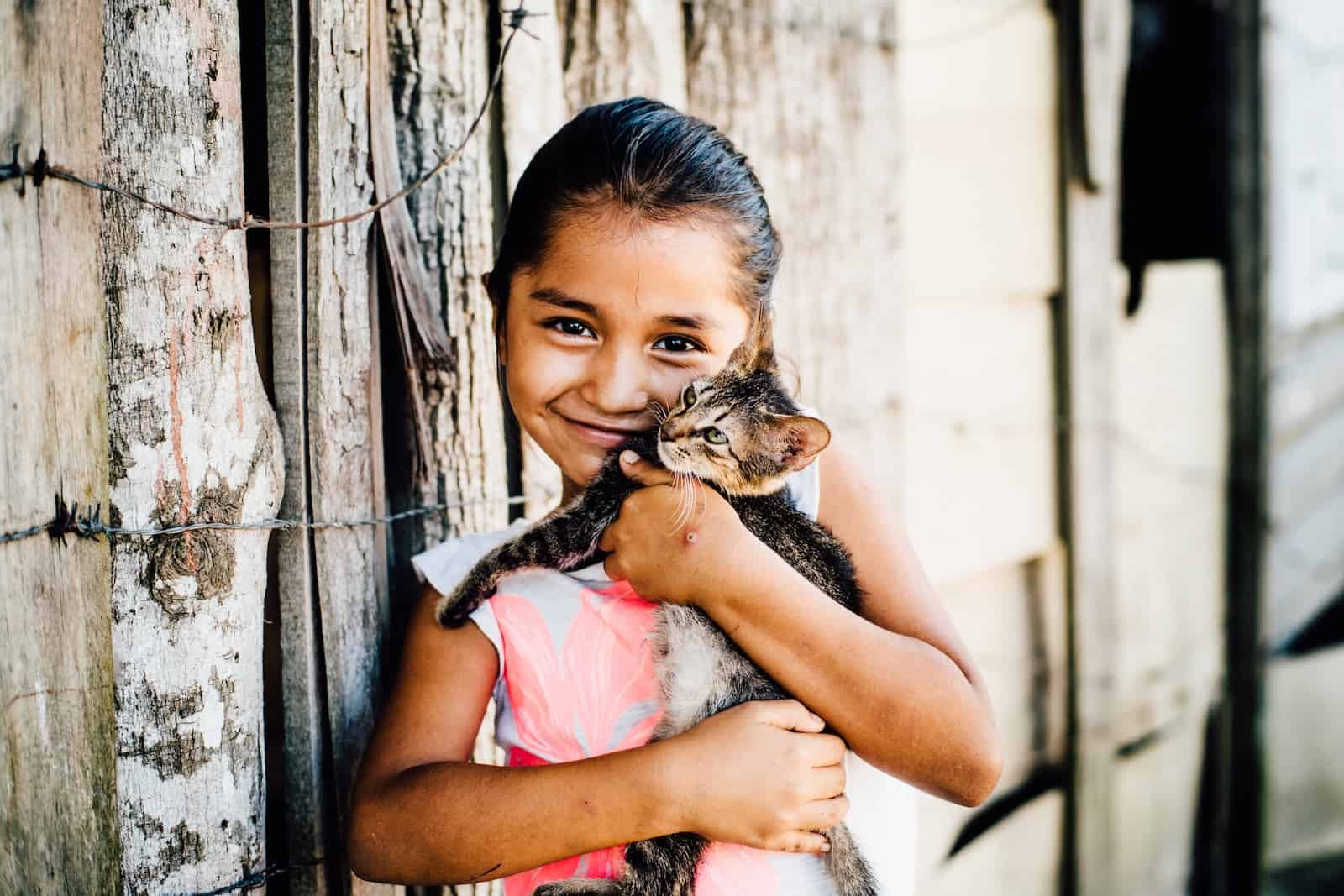 A young girl wearing a white T-shirt cuddles a kitten in front of a wooden fence.