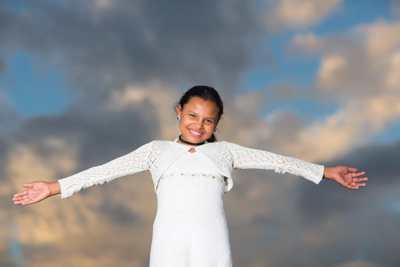 A girl in a white dress holds her arms out to the side, in front of a beautiful sky. 