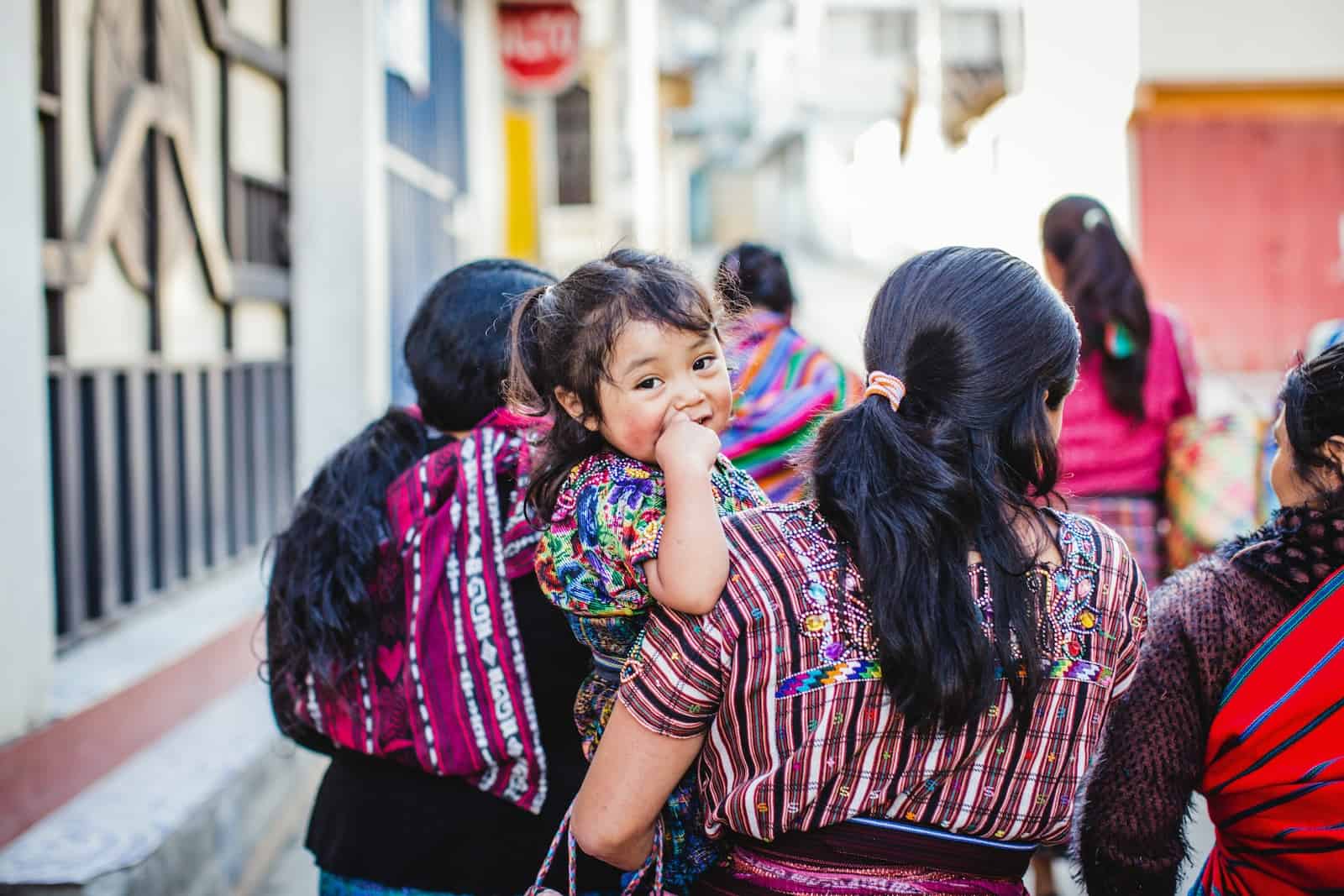 A baby is held by her mom, surrounded by other women walking down a street, wearing colorful clothing.