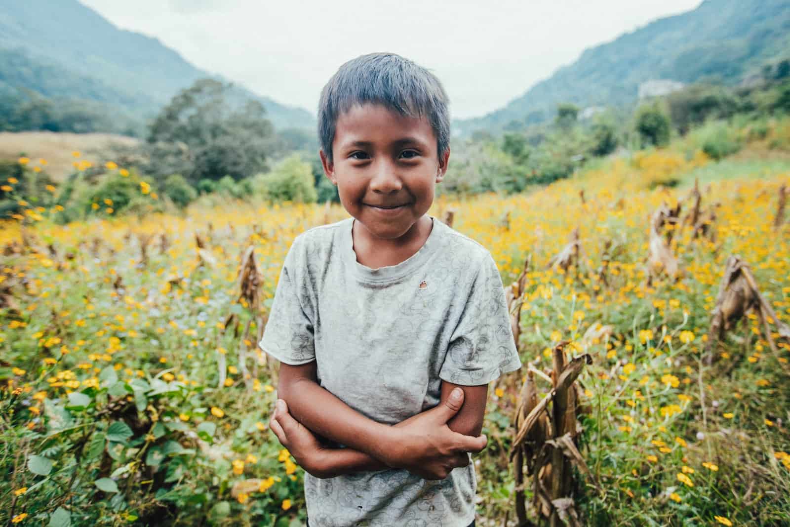 A young boy in a gray T-shirt stands with his arms crossed in front of a field of yellow flowers. 