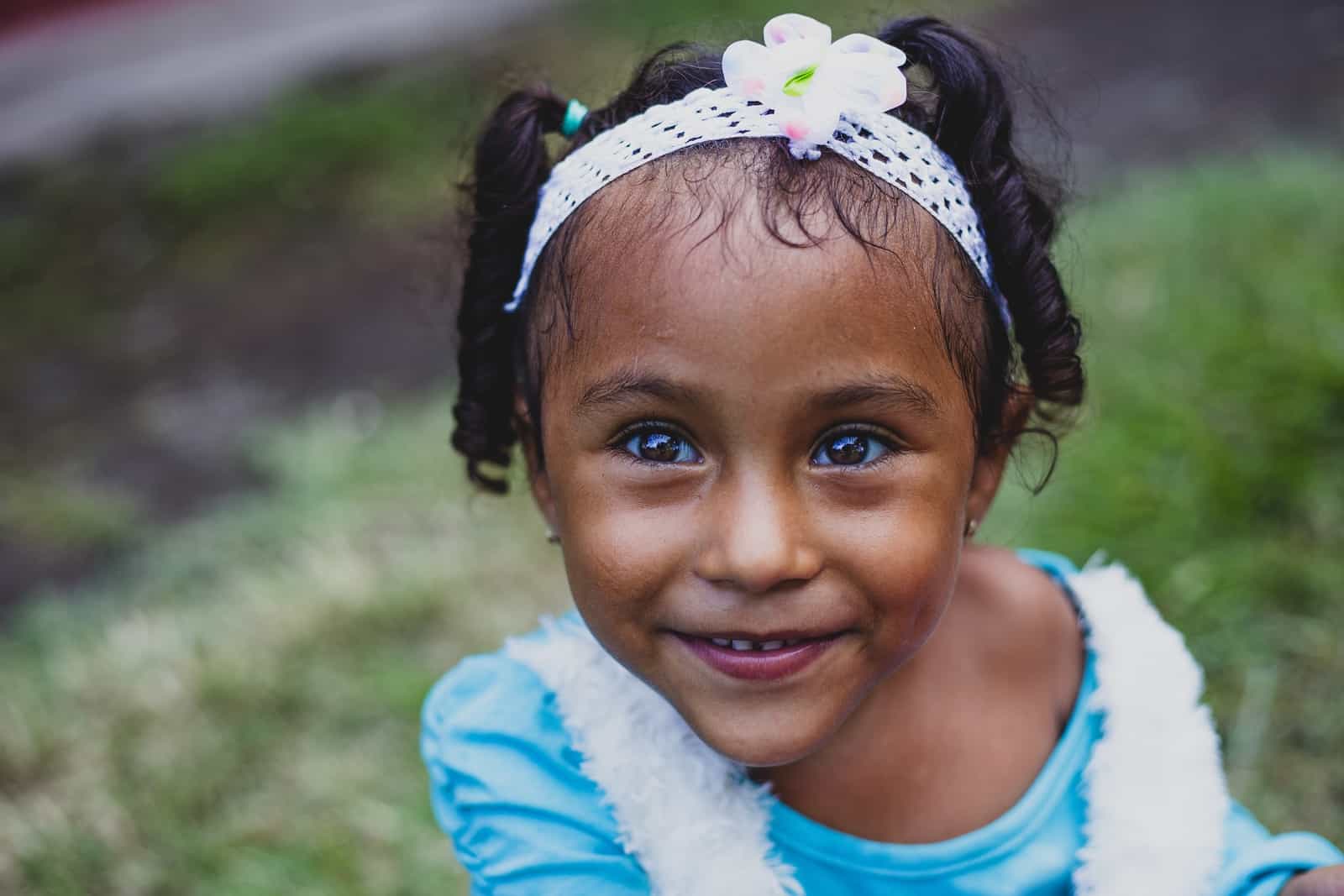 A girl in a blue shirt and white headband with a flower on it looks up into the camera. 