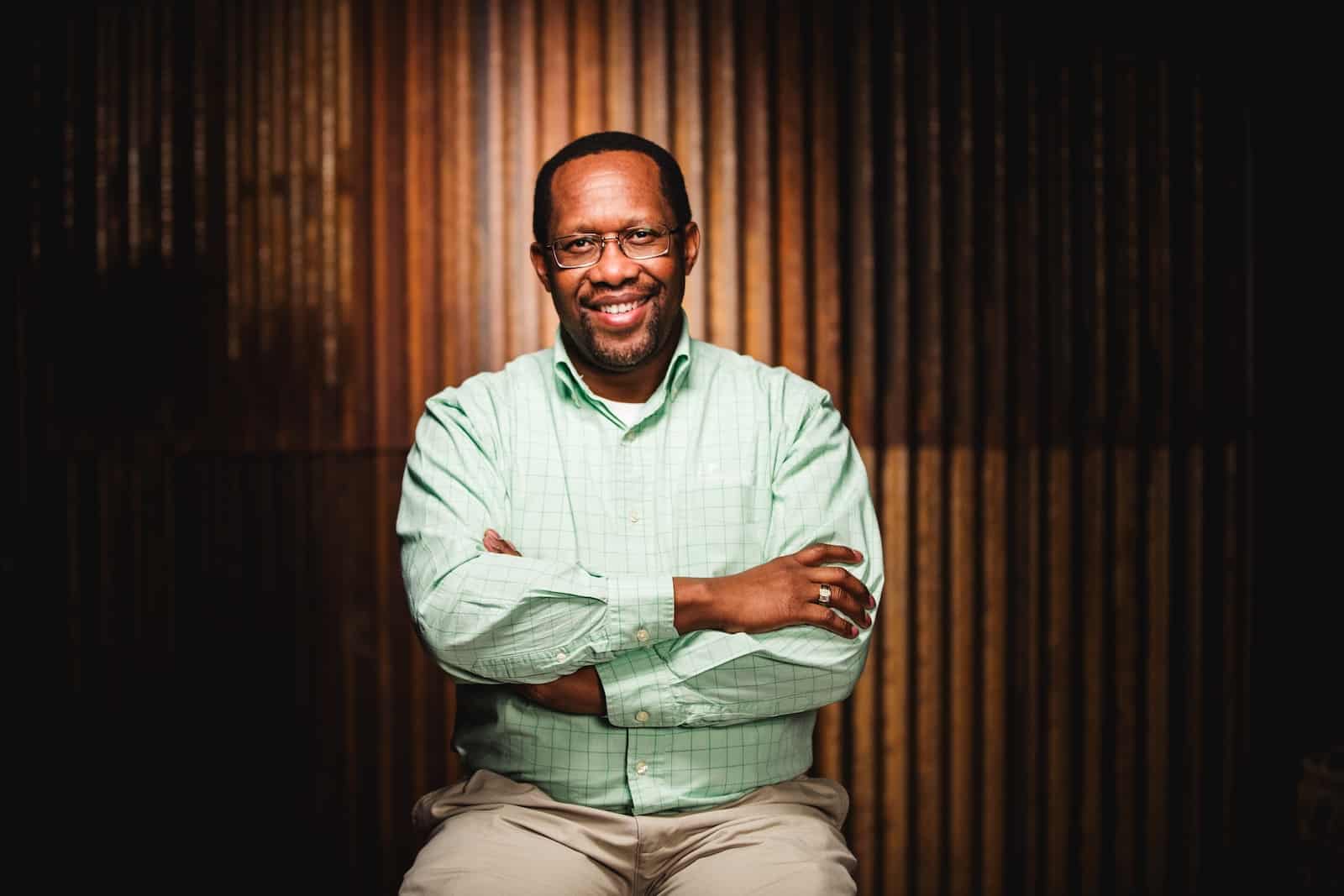 A man in a green button-up shirt and khaki pants sits on a stool, arms crossed, in front of a corrugated metal background.