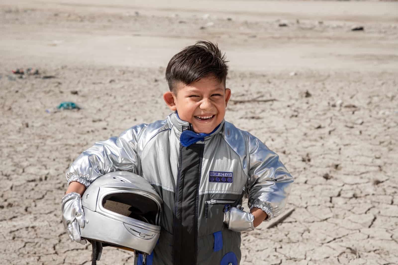 A boy wearing a silver suit and holding a silver helmet stands with one hand on his hip, smiling, in front of a dry, parched dirt background. 