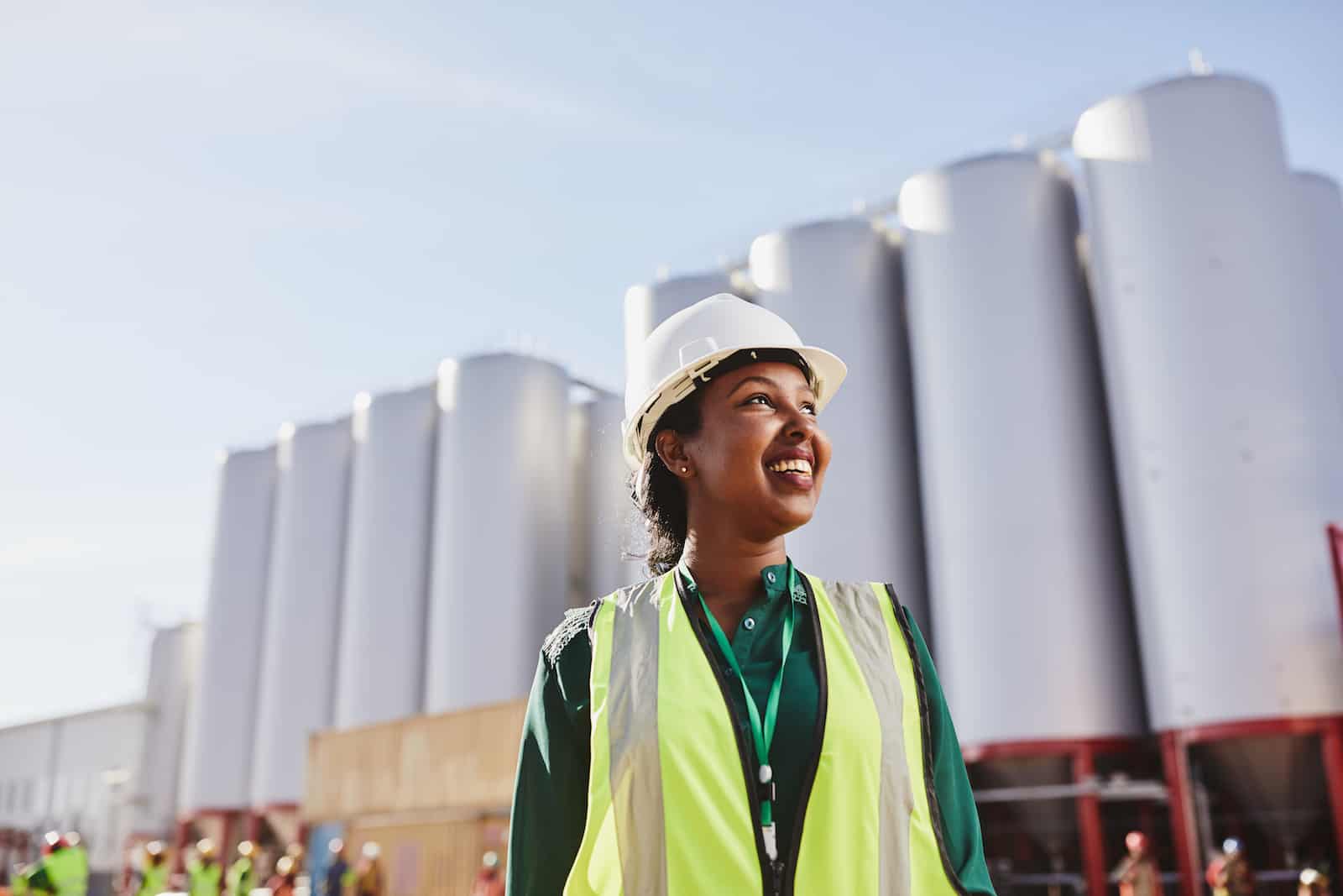 A woman in a yellow safety vest and hard hat stands in front of a manufacturing plant, looking up to the sky. 
