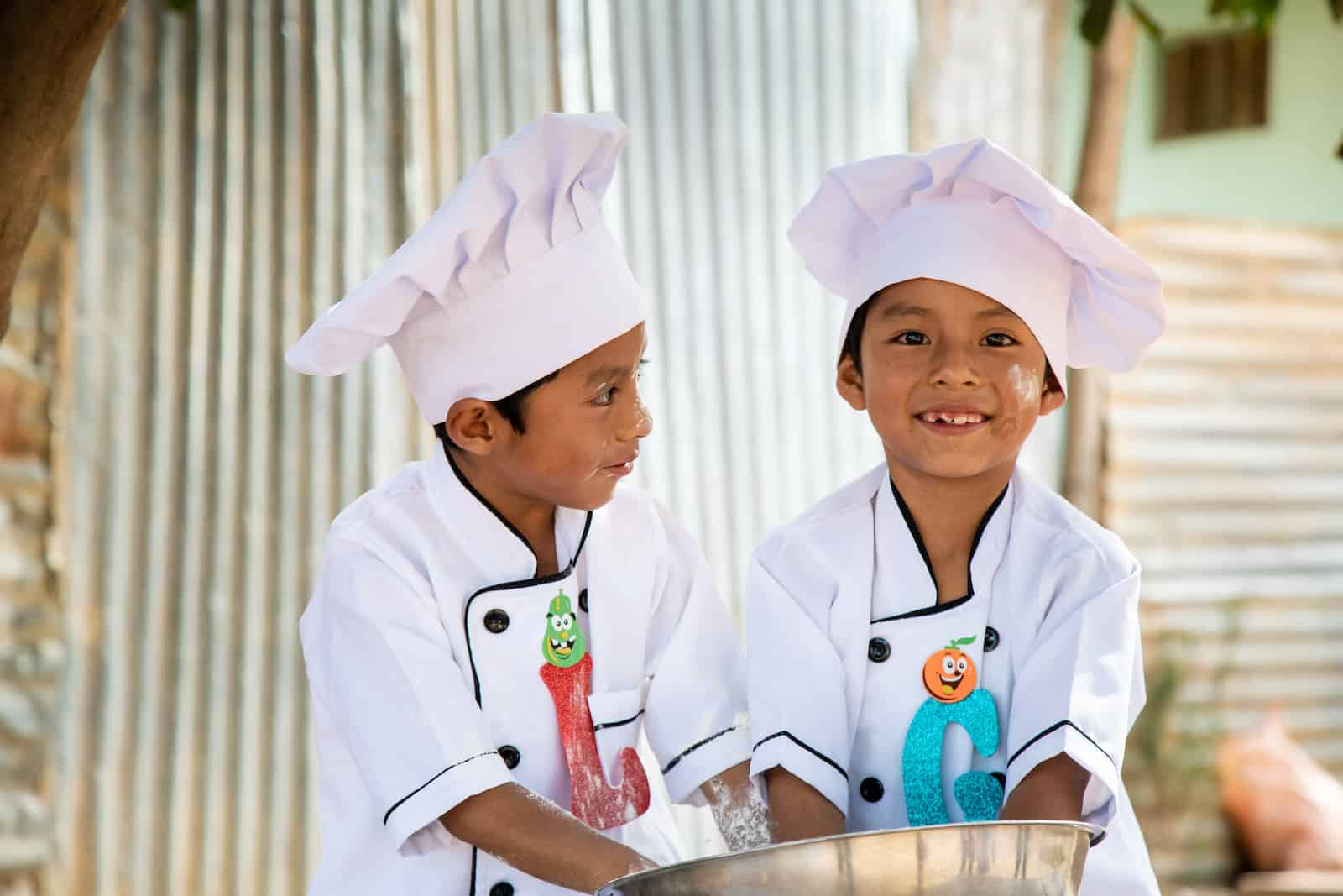 Two boys wearing chefs hats and white chefs jackets stand at a table, with their hands in a large silver bowl. One smiles at the camera and one looks at the other boy. 