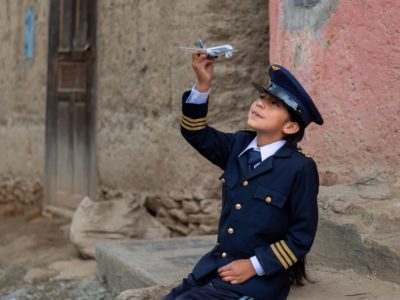 A girl wearing a navy blue pilot's uniform and cap sits on a sidewalk, holding a toy airplane up into the sky, looking at it.