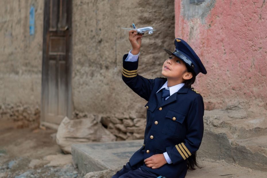 A girl wearing a navy blue pilot's uniform and cap sits on a sidewalk, holding a toy airplane up into the sky, looking at it.
