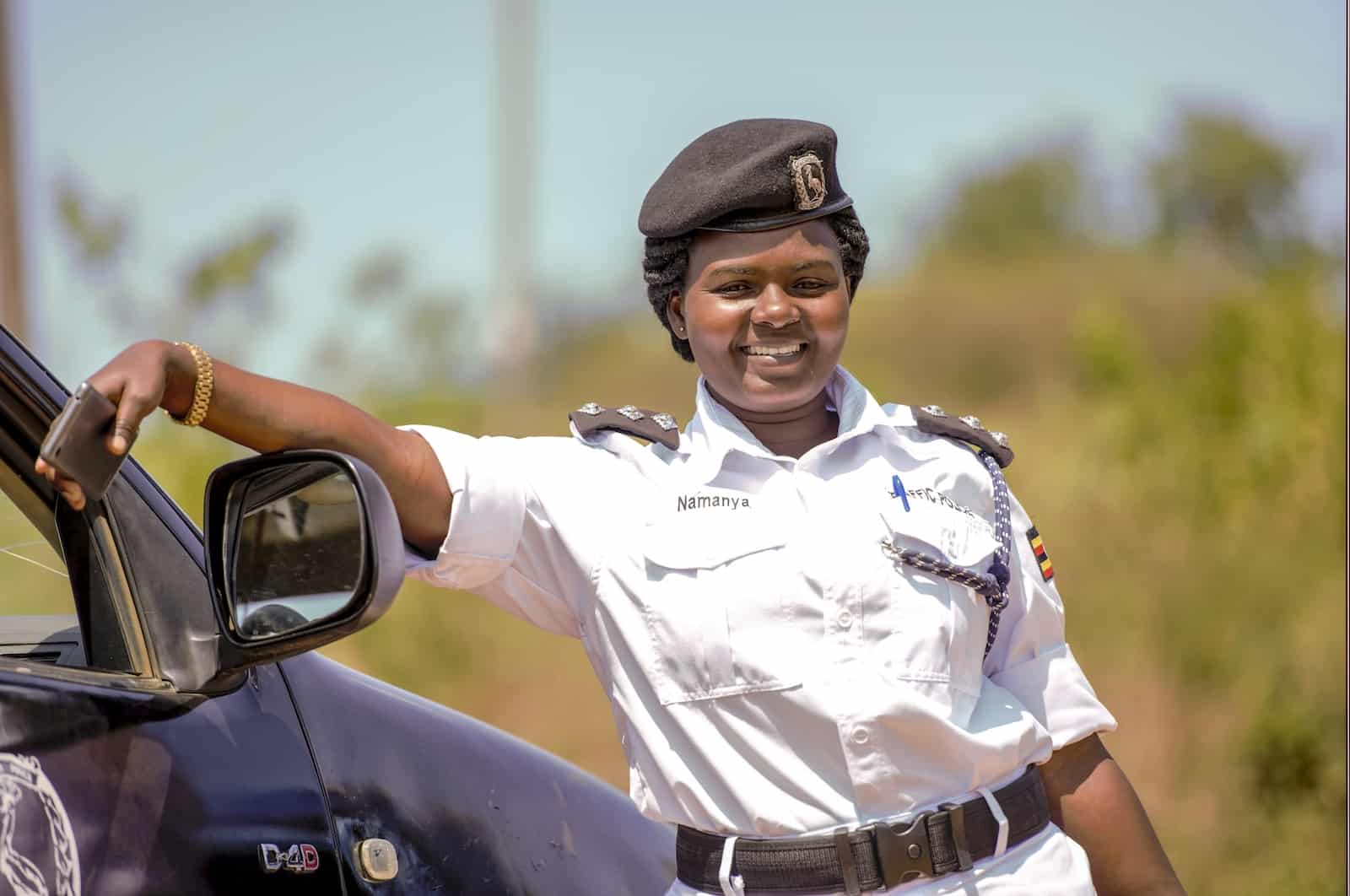 A woman wearing a white police uniform and black cap leans against a black car. 