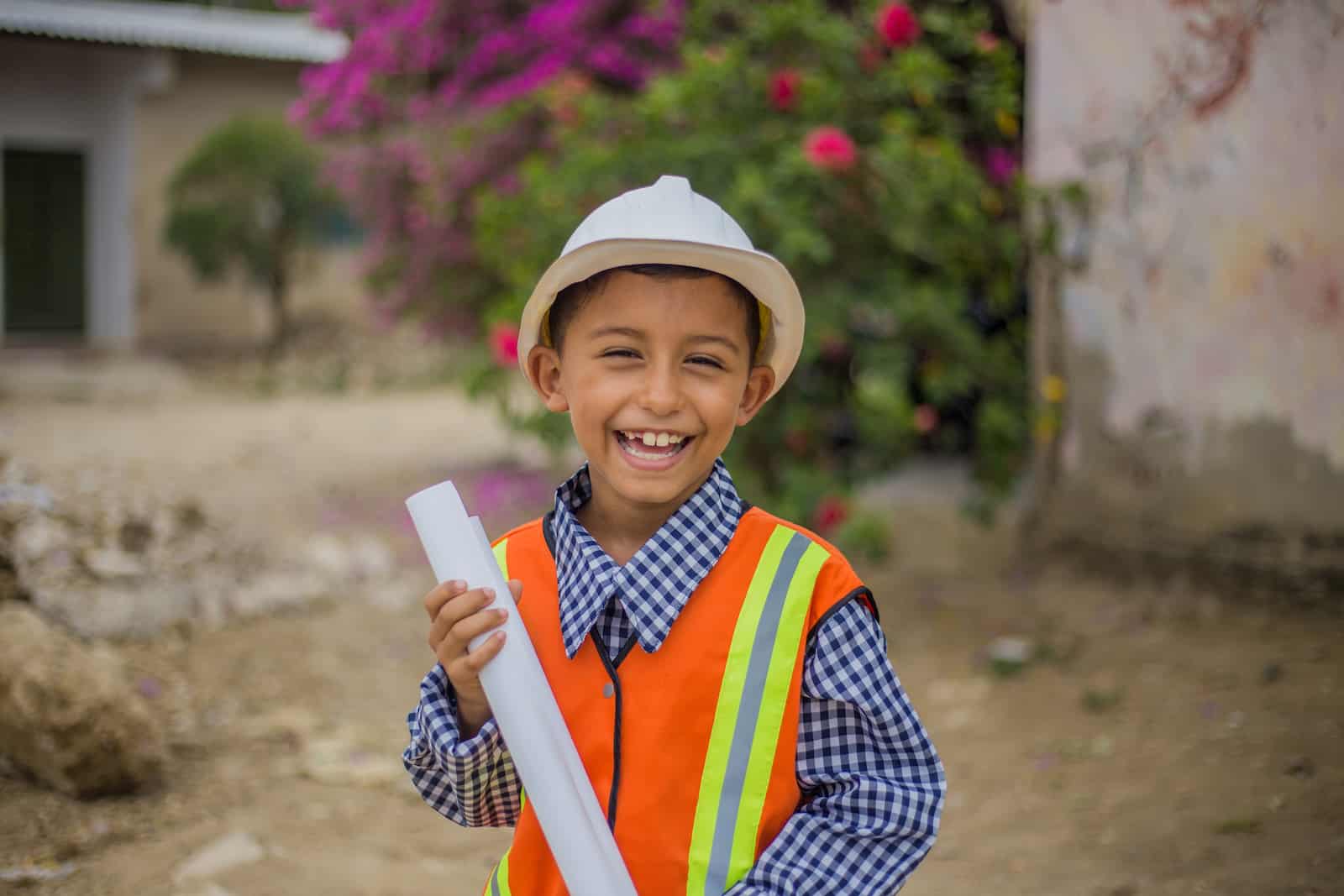 A boy wears a orange safety vest over a blue shirt, along with a hard hat. He holds a rolled up piece of paper, smiling at the camera. He stands on a street in front of a bush with pink flowers. 