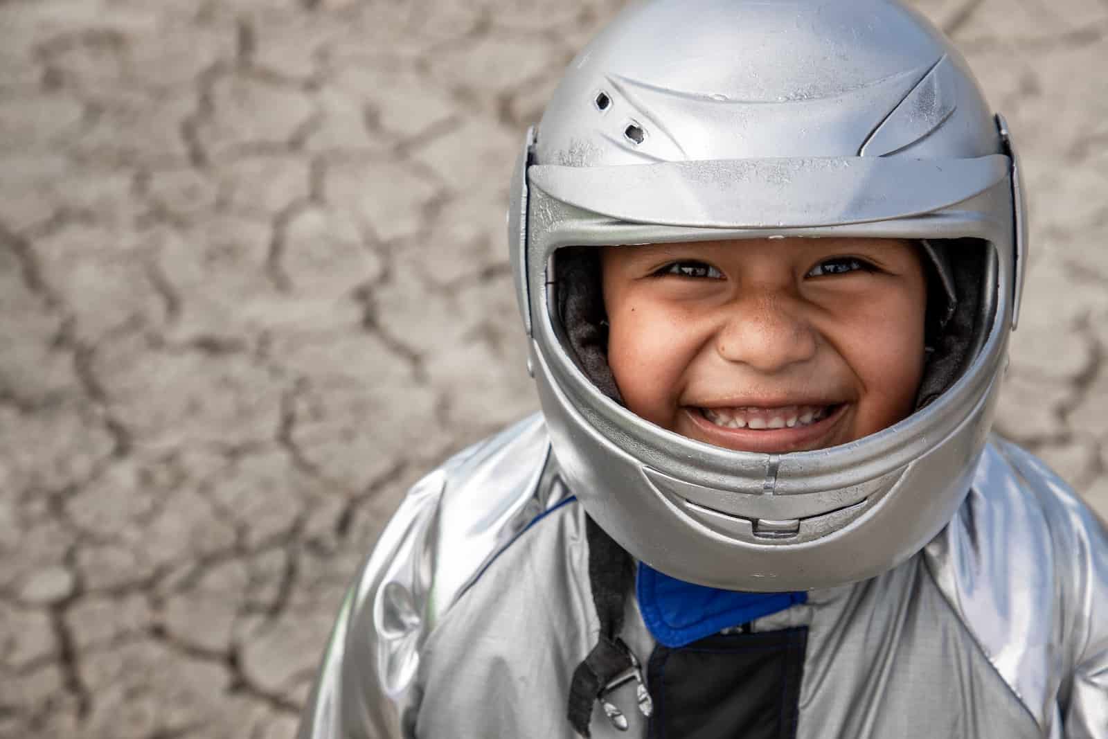 A boy wearing a silver helmet and silver astronaut suit smiles up at the camera. He stands in front of dry, parched ground. 