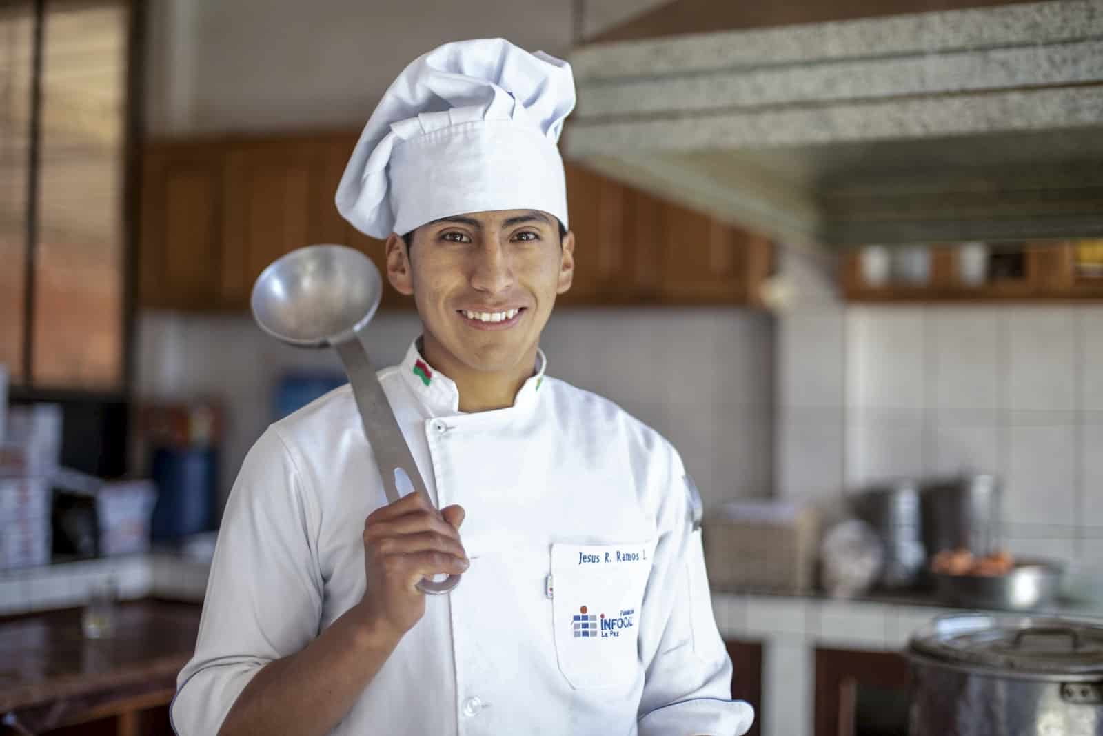 A man wearing a chef's hat and white chef's coat stands in a kitchen, holding a large ladle over his shoulder, smiling at the camera. 