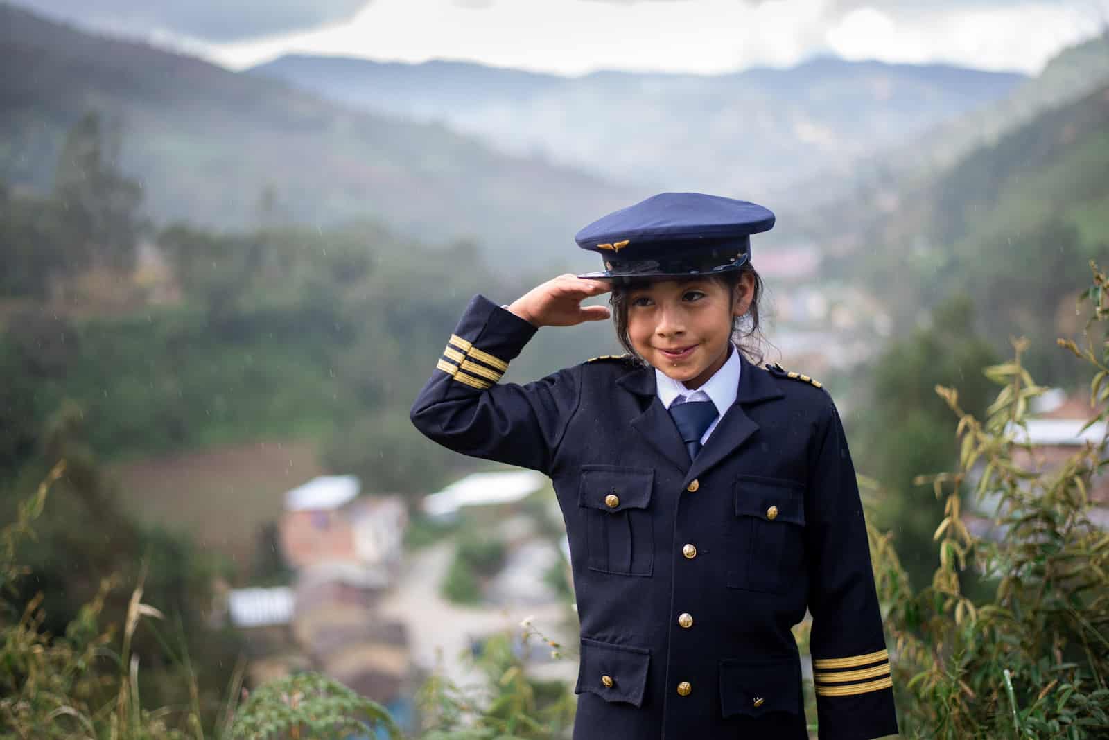 A girl in a blue pilot's uniform and hat salutes. Behind her is a village in a mountain valley. 