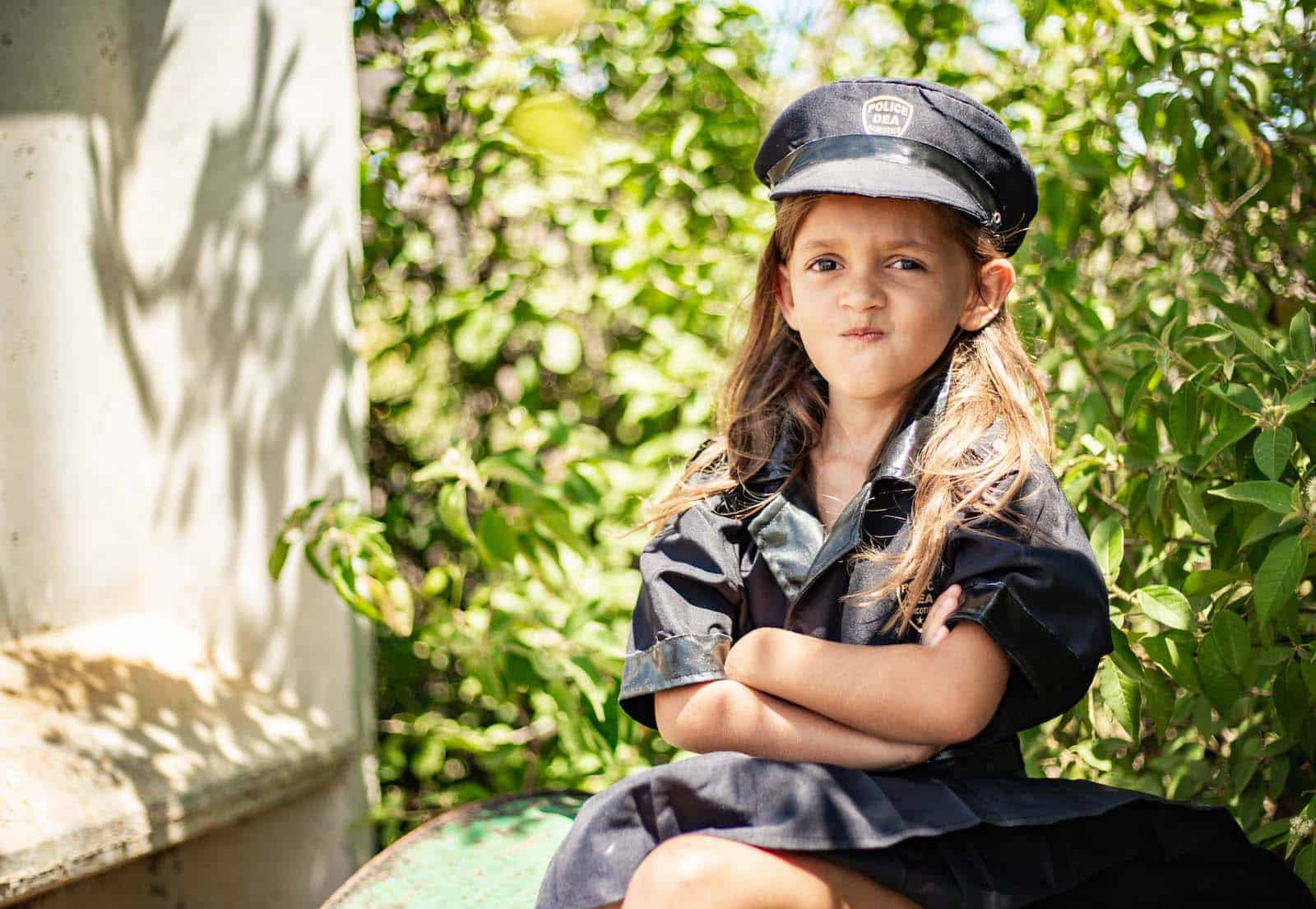 A young girl wearing a black police officer costume and cap crosses her arms in front of her, while sitting in front of a bush. Her lips are pursed in a funny face. 