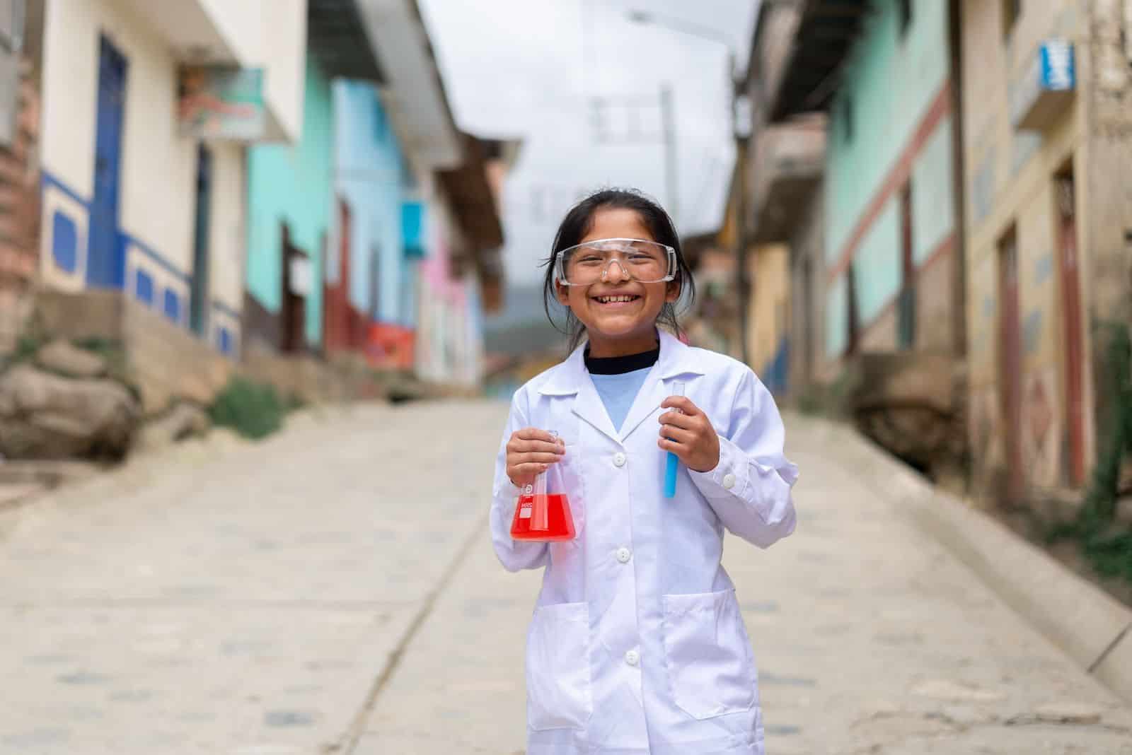 A girl wearing a white lab coat and safety glasses holds two beakers full of red and blue liquid, smiling at the camera. She stands on a street with a row of concrete homes. 