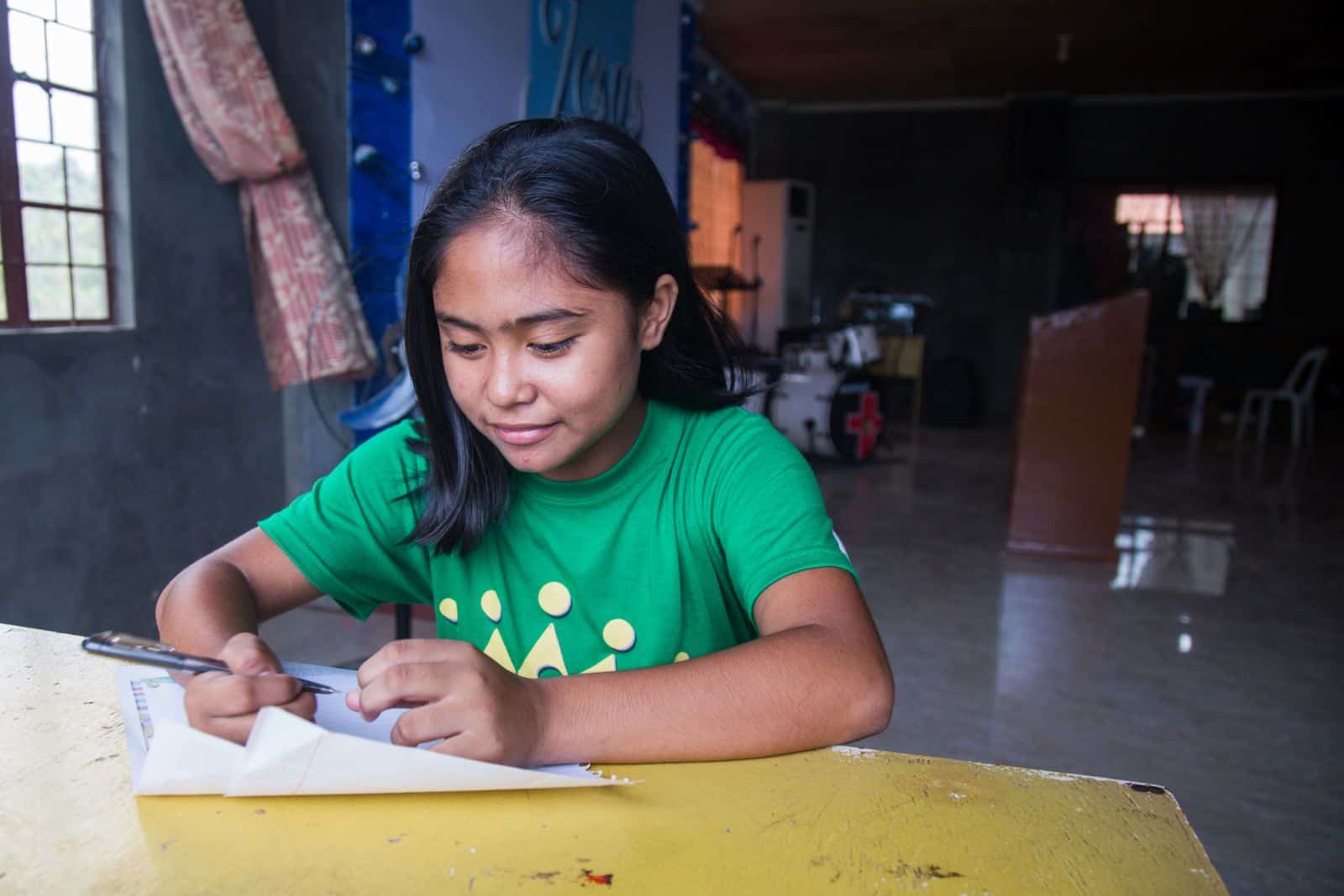 A girl in a green T-shirt sits at a table inside of a church, writing a letter.