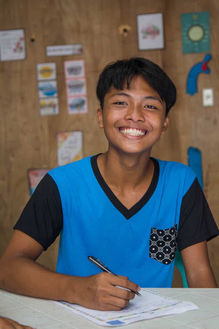 A boy in a blue and black T-shirt smiles widely, sitting at a desk writing a letter. 