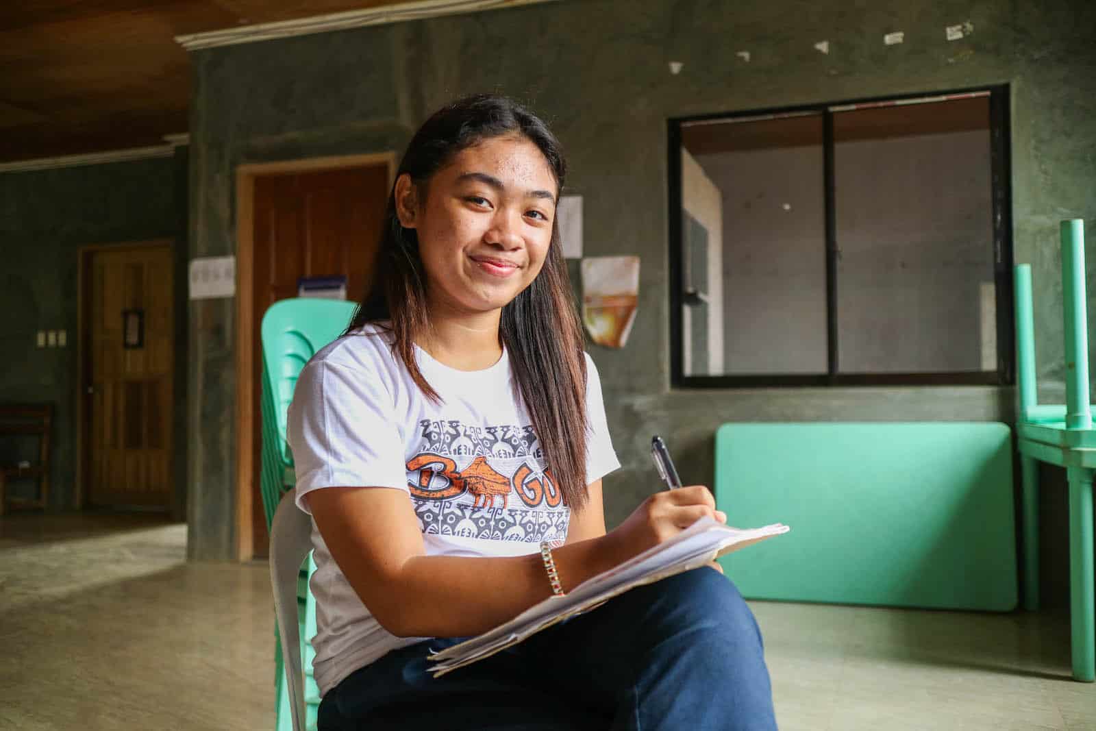 A girl in a white T-shirt and jeans sits at a chair, with a pen and paper in her lap, smiling at the camera.