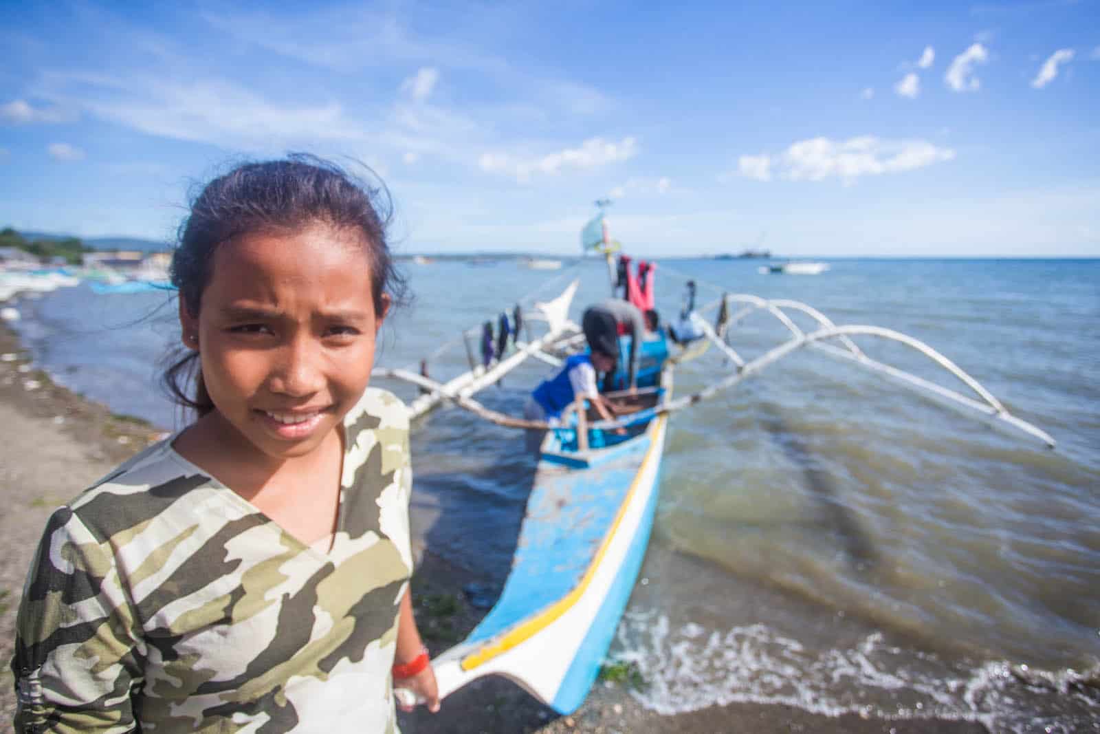 A girl in a camo shirt stands on the beach in front of a long blue canoe with men on it. 