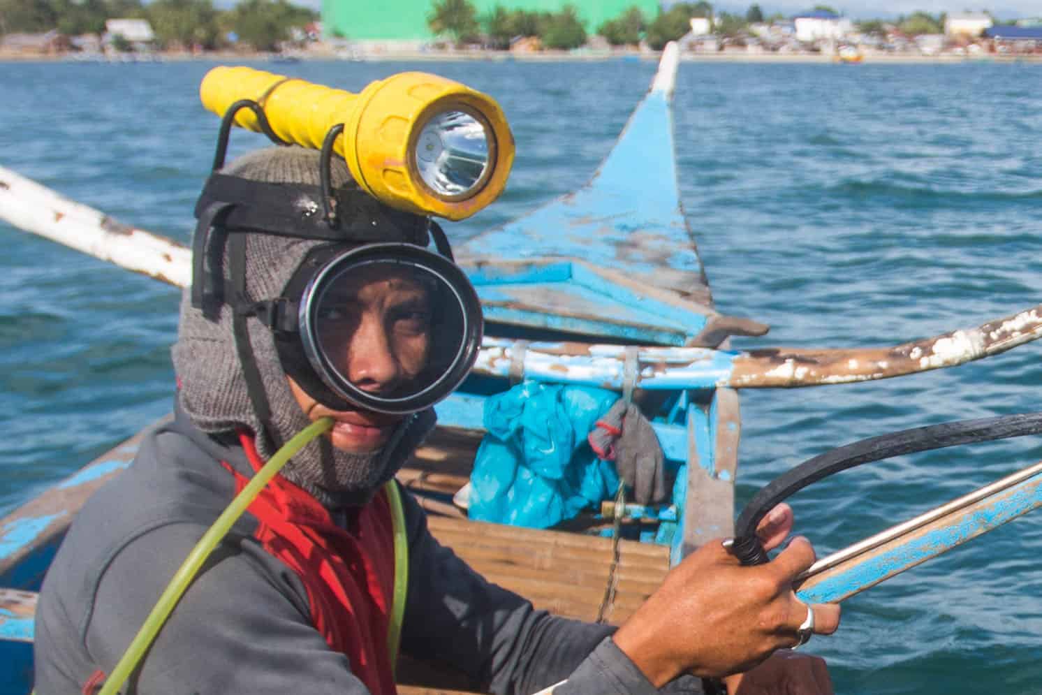 A man sits on a boat, with a swim mask on, a flashlight strapped to his head and a plastic tube in his mouth, ready to go pa-aling, one of the most dangerous jobs.