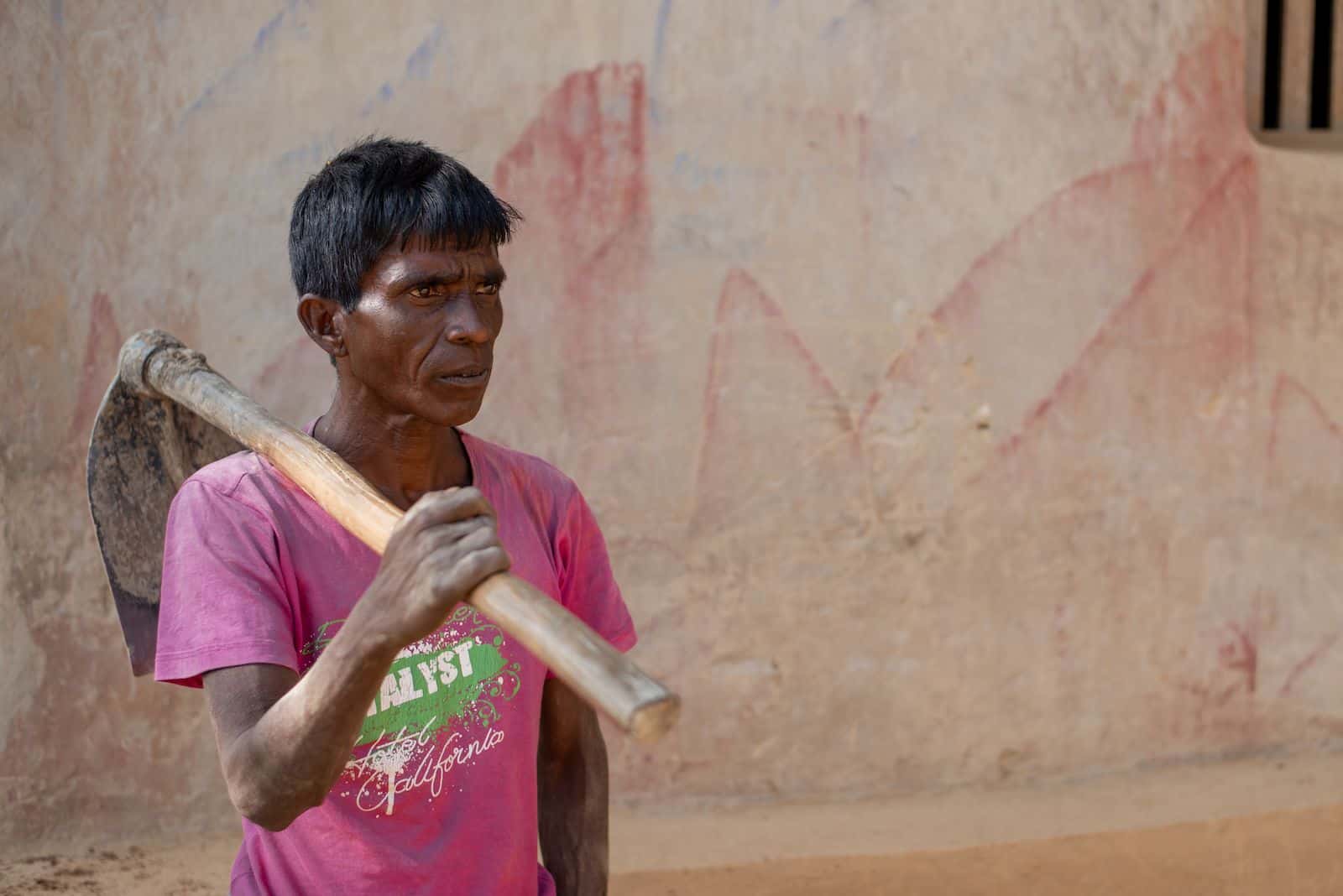 A man wearing a pink shirt stands with a hoe on his shoulder, looking stoicly to the side.