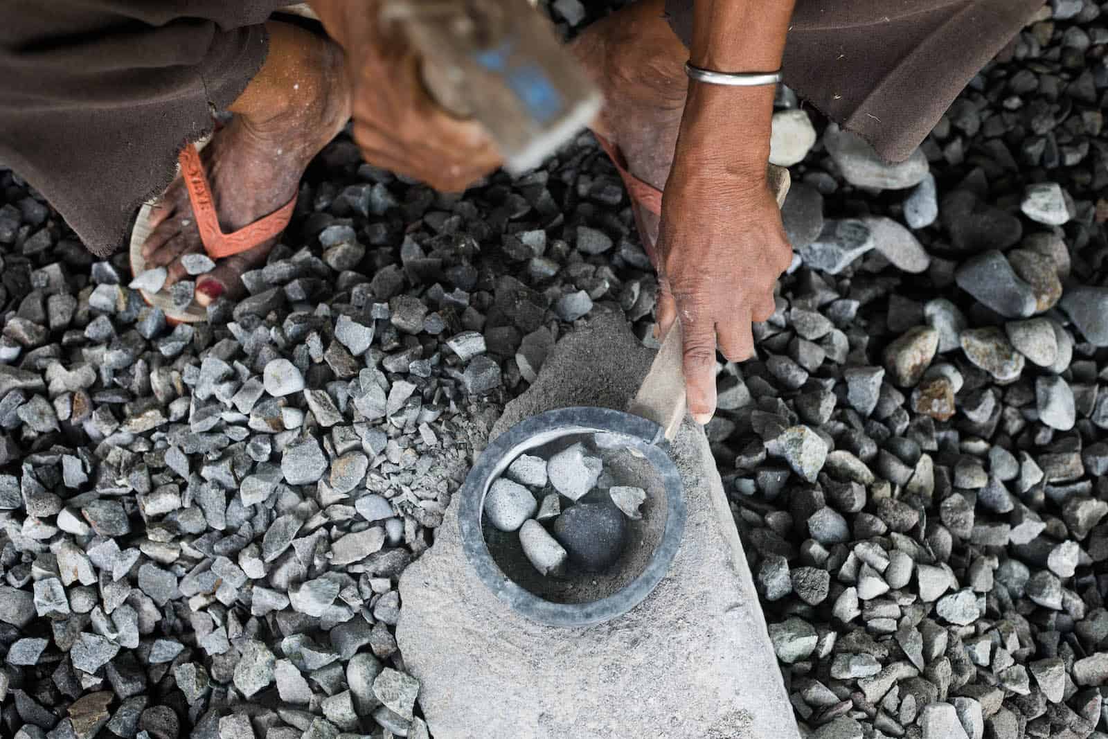 A hand holds a hammer, poised over a group of pebbles on a stone. 