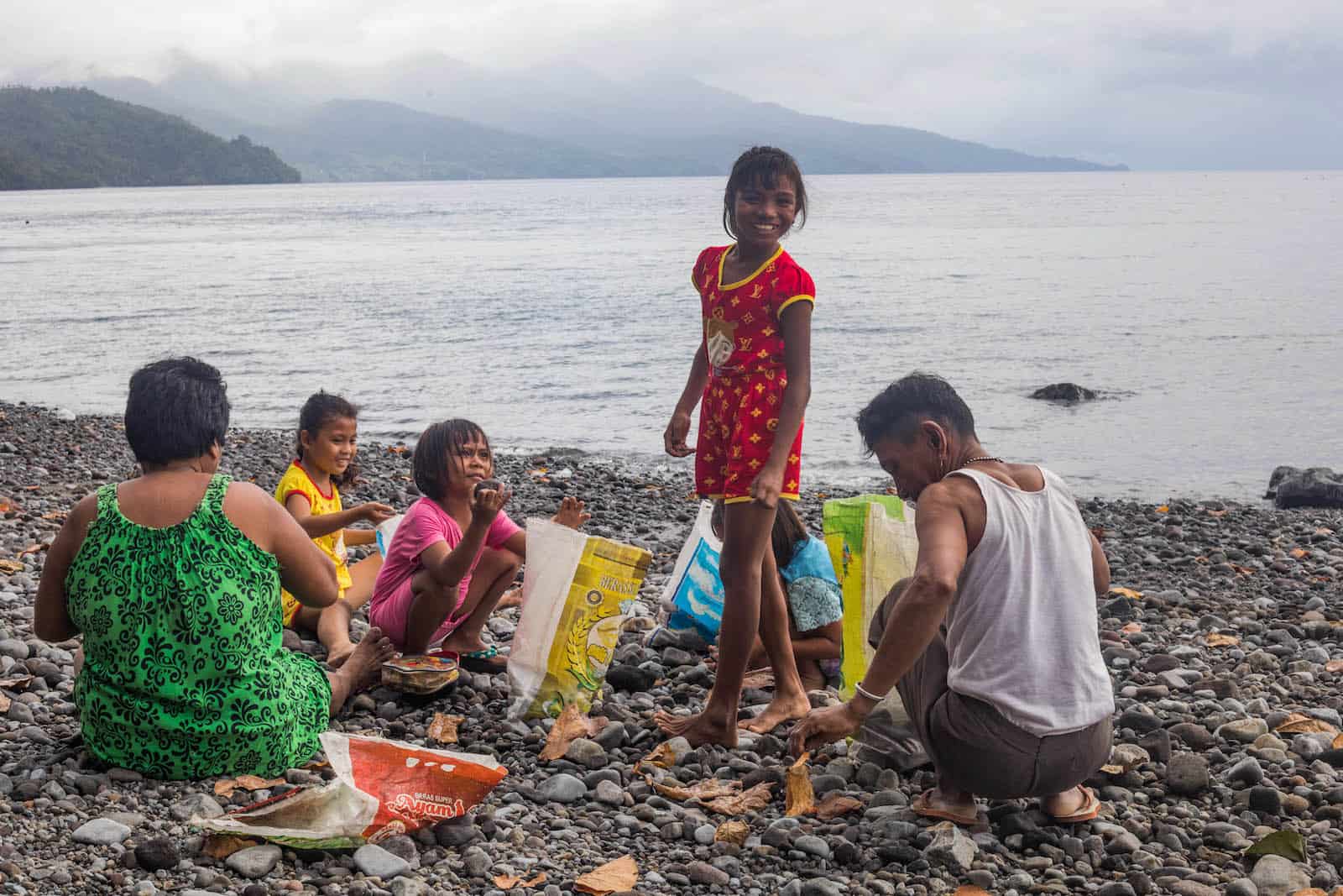 A girl stands on the beach, surrounded by girls and her grandparents collecting rocks. 