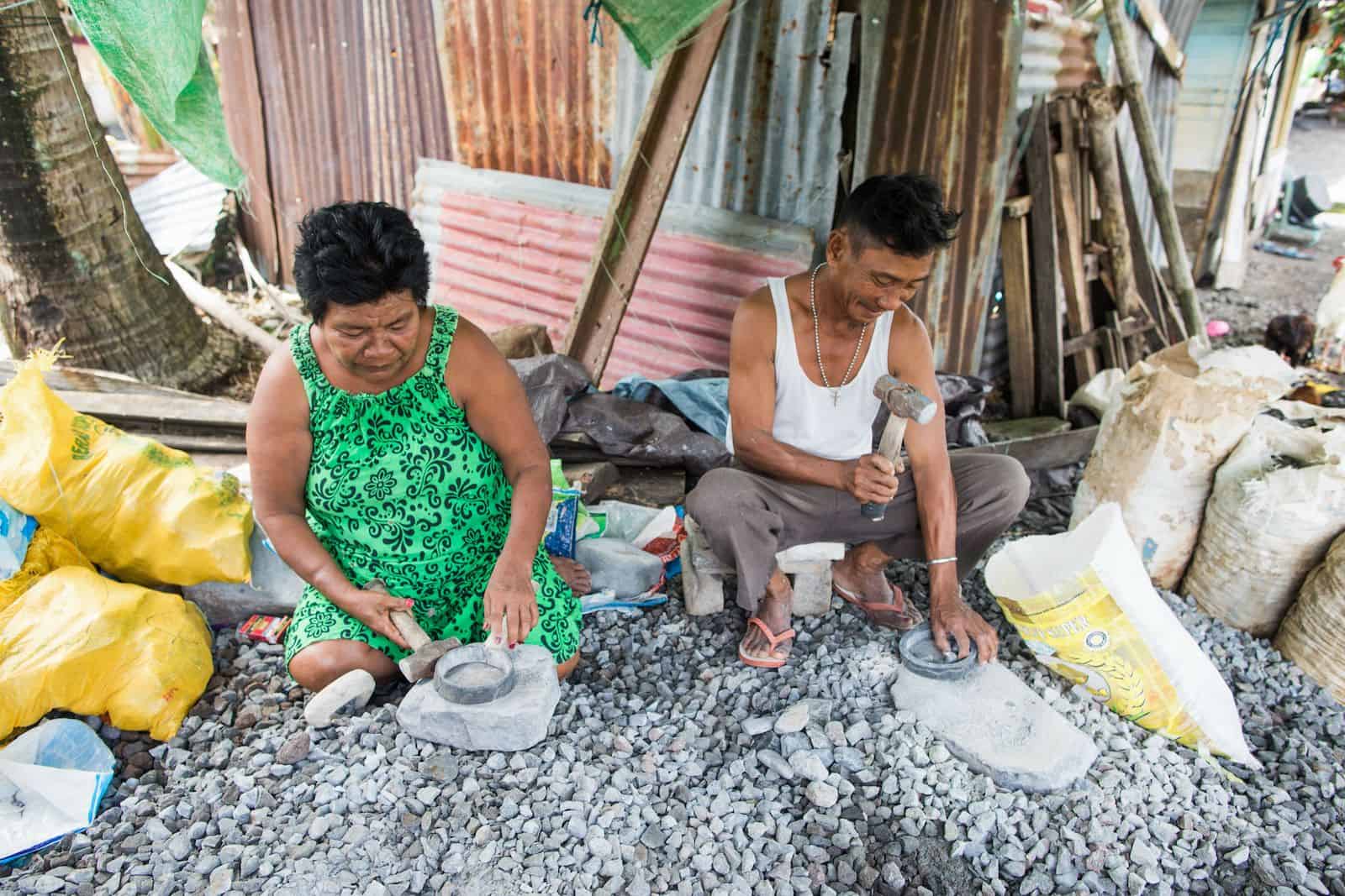 A man and woman crouch on the ground breaking rocks with small hammers. 