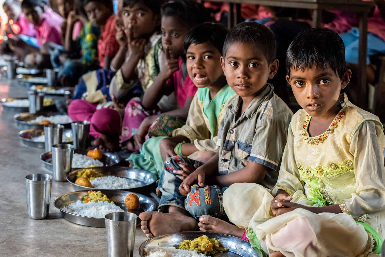 A group of children sit on the floor in a line, eating plates of rice. Report: Hunger Is on the Rise in the World