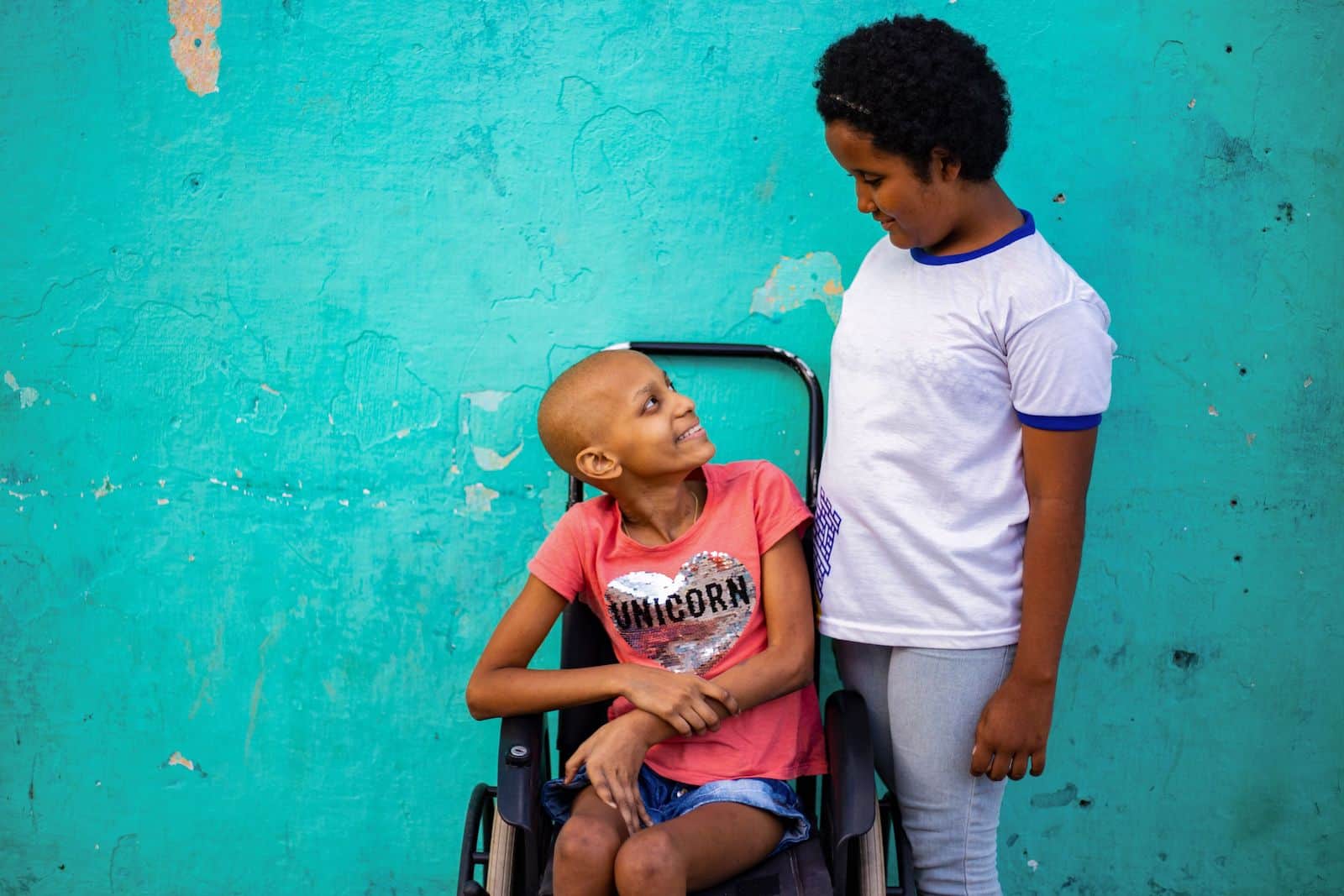 A girl in a white T-shirt stands next to a girl in a pink shirt, sitting in a wheelchair. They are looking at each other in front of a turquoise wall. 