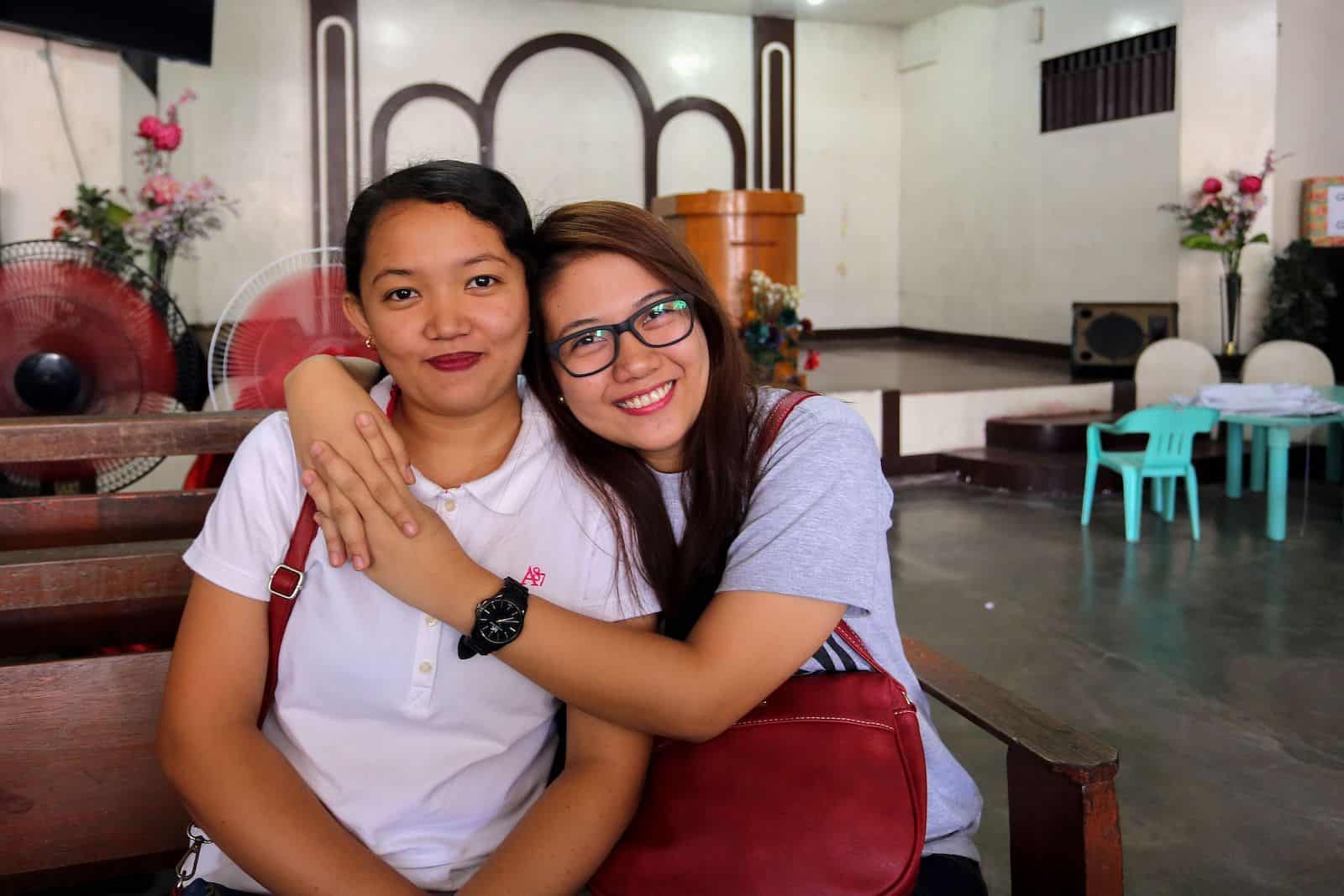 Two young women sit inside a church, one with her arms around the other, both smiling. 