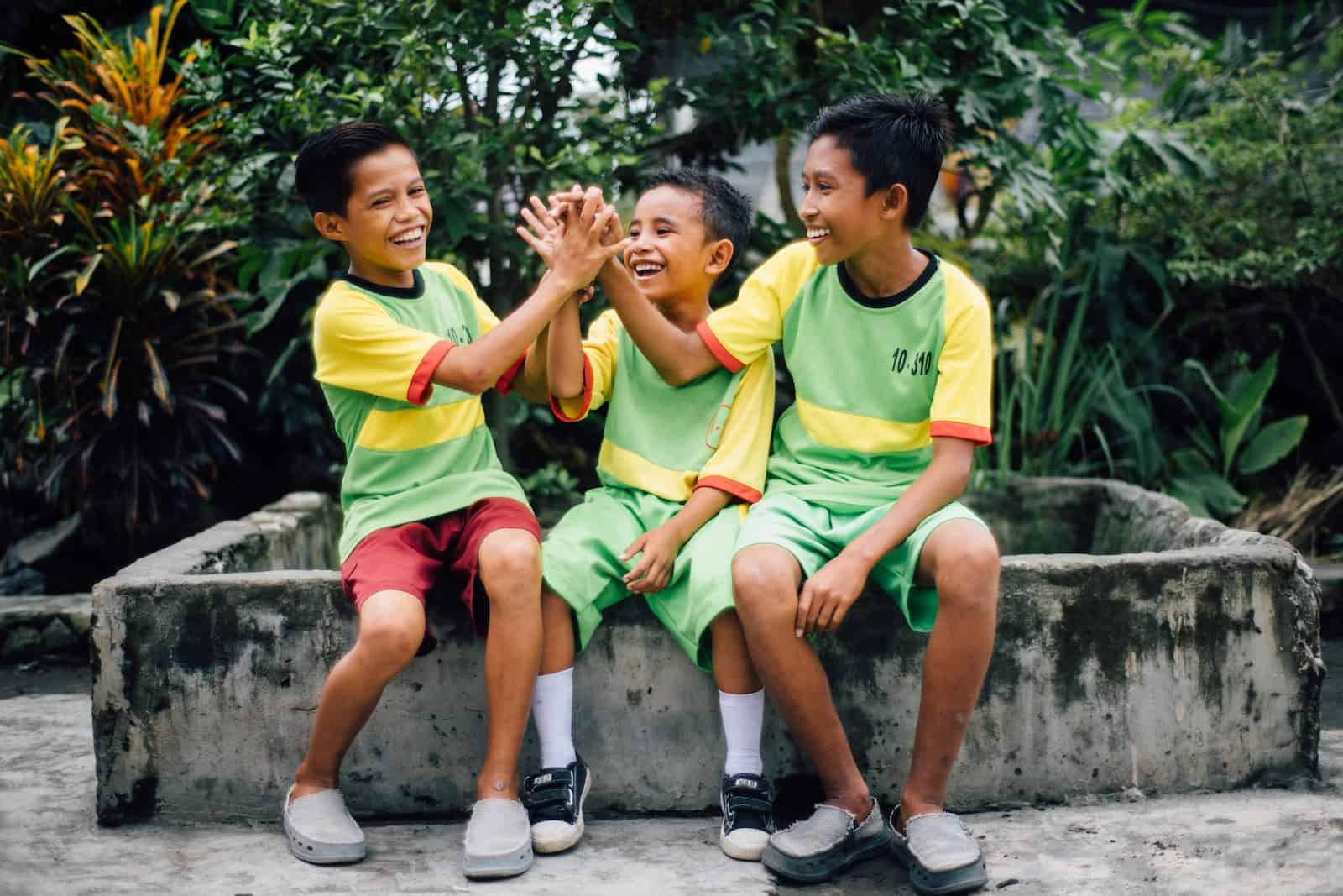 International Day of Friendship: Three boys sit outside wearing green and yellow sports uniforms, giving each other high fives. 