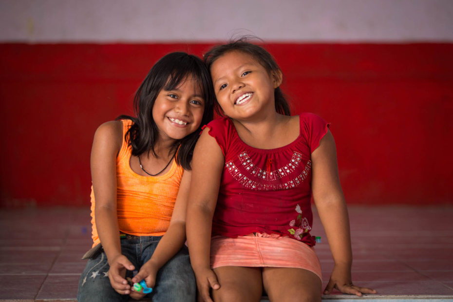 Two young girls sit inside on a step in front of a red wall, smiling.