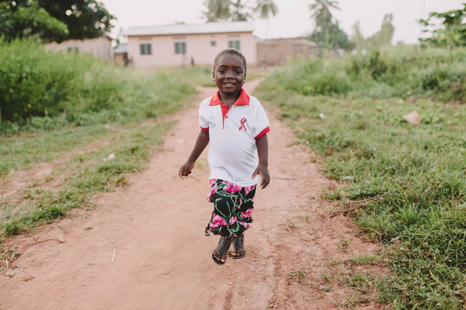 A girl in a white T-shirt and patterned skirt runs down a dirt path, smiling, with a house in the background.