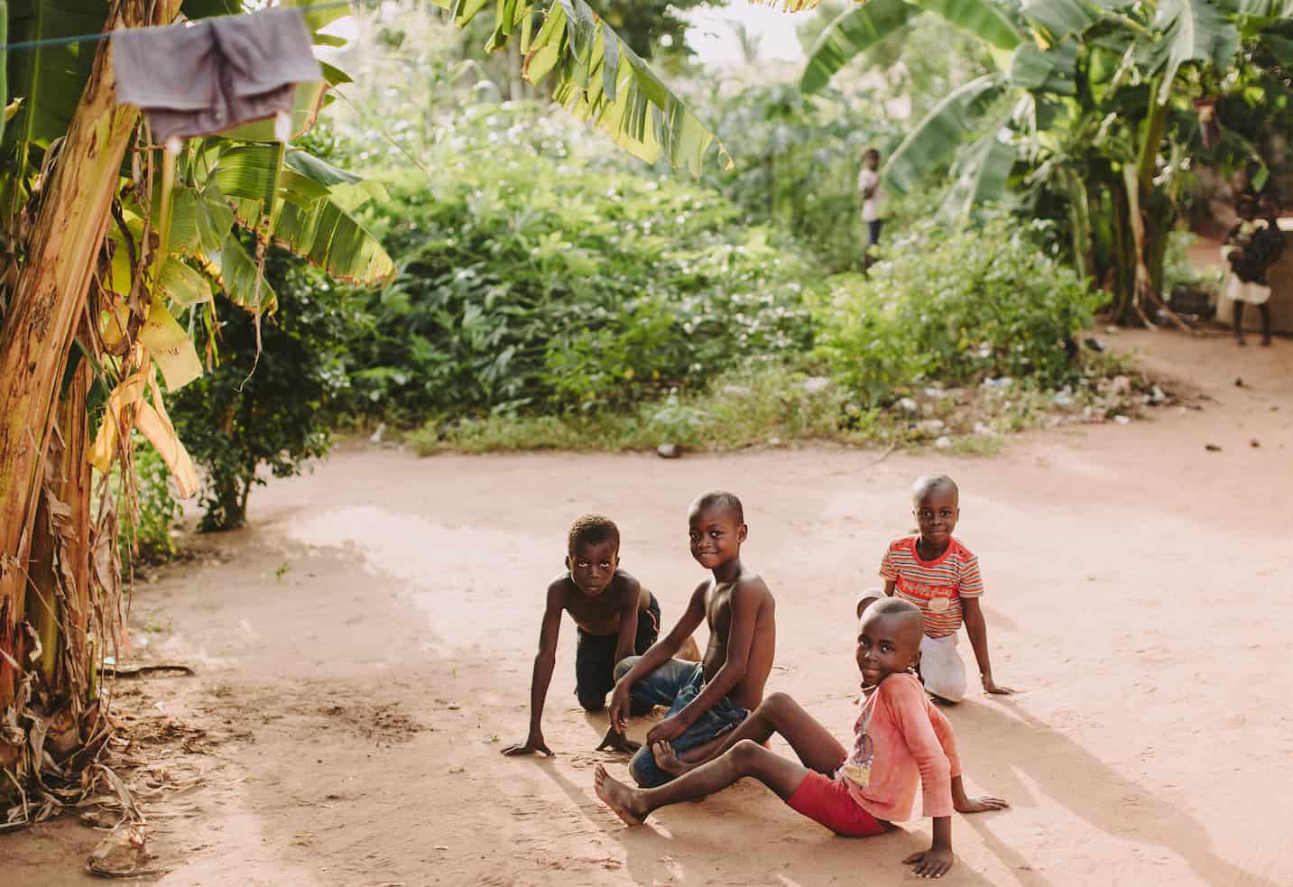 Children sit on the dirt ground, playing. There are palm trees in the background.