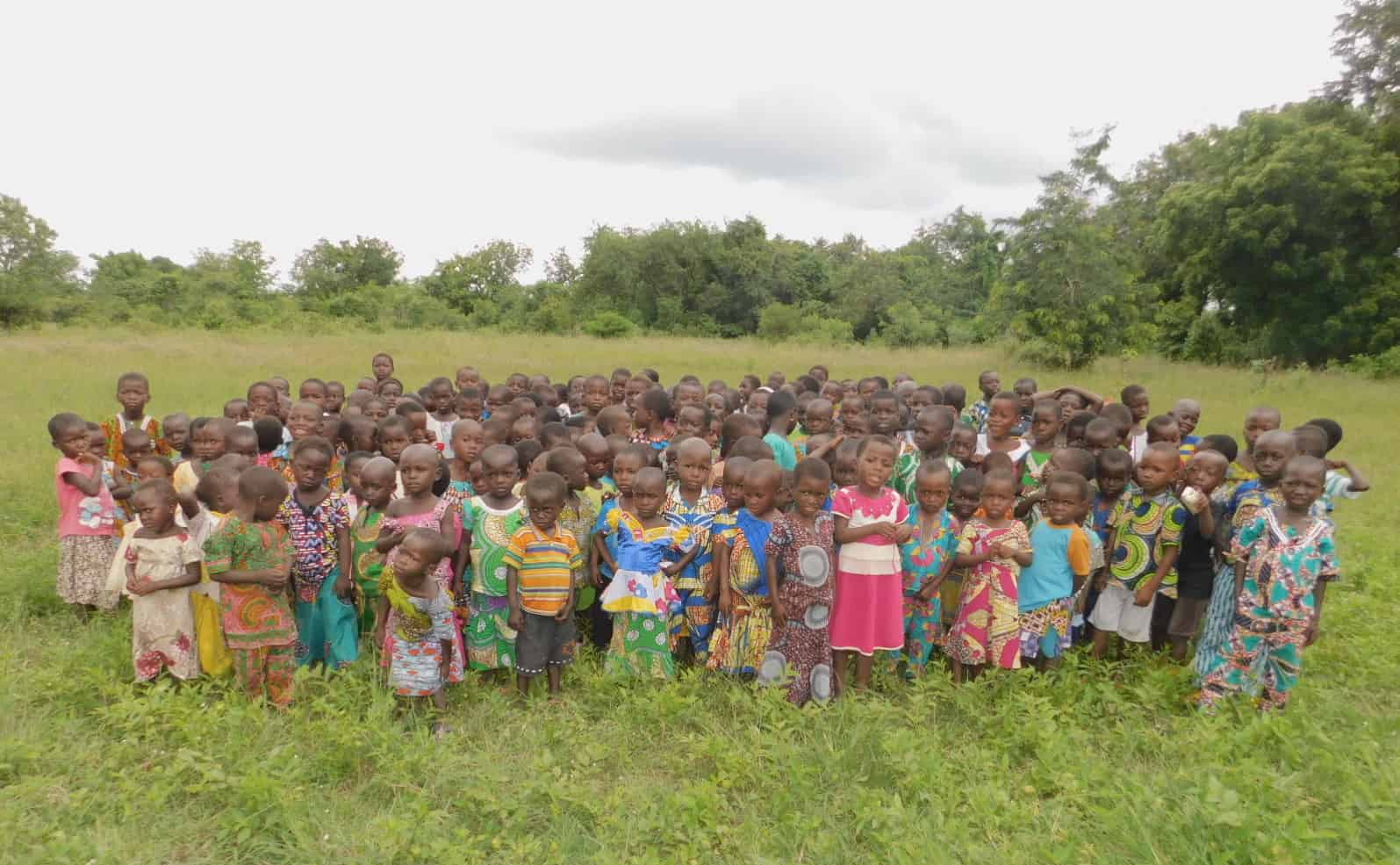 A large group of children in colorful clothes stand in a field in rural Togo.