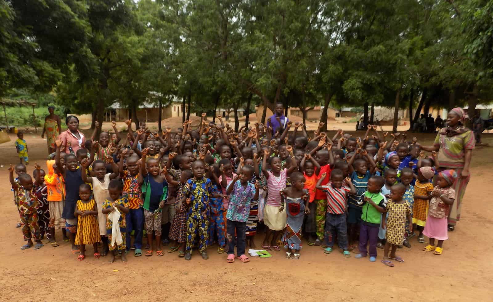 A large group of children in colorful clothes stands outside in front of a row of trees in rural Togo.