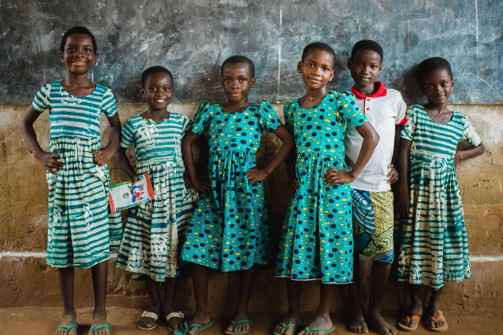 A group of girls in green patterned dresses stands in front of a chalkboard in a classroom, with hands on hips. 