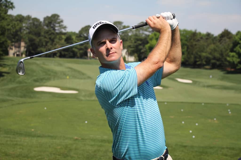 A man, Kevin Streelman, in a white baseball cap and blue shirt swings golf club, standing in front of a golf course. 