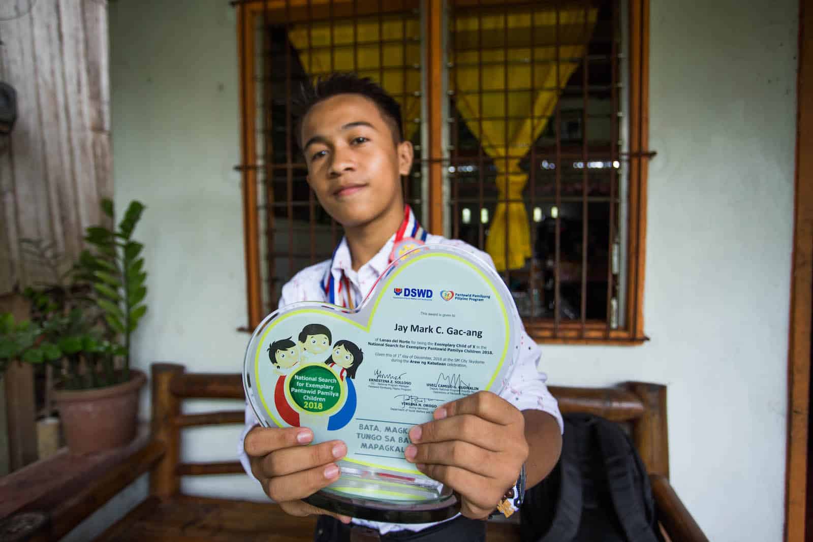 A teen boy stands outside a window, holding an award plaque for the Most Excellent Child in the Philippines.