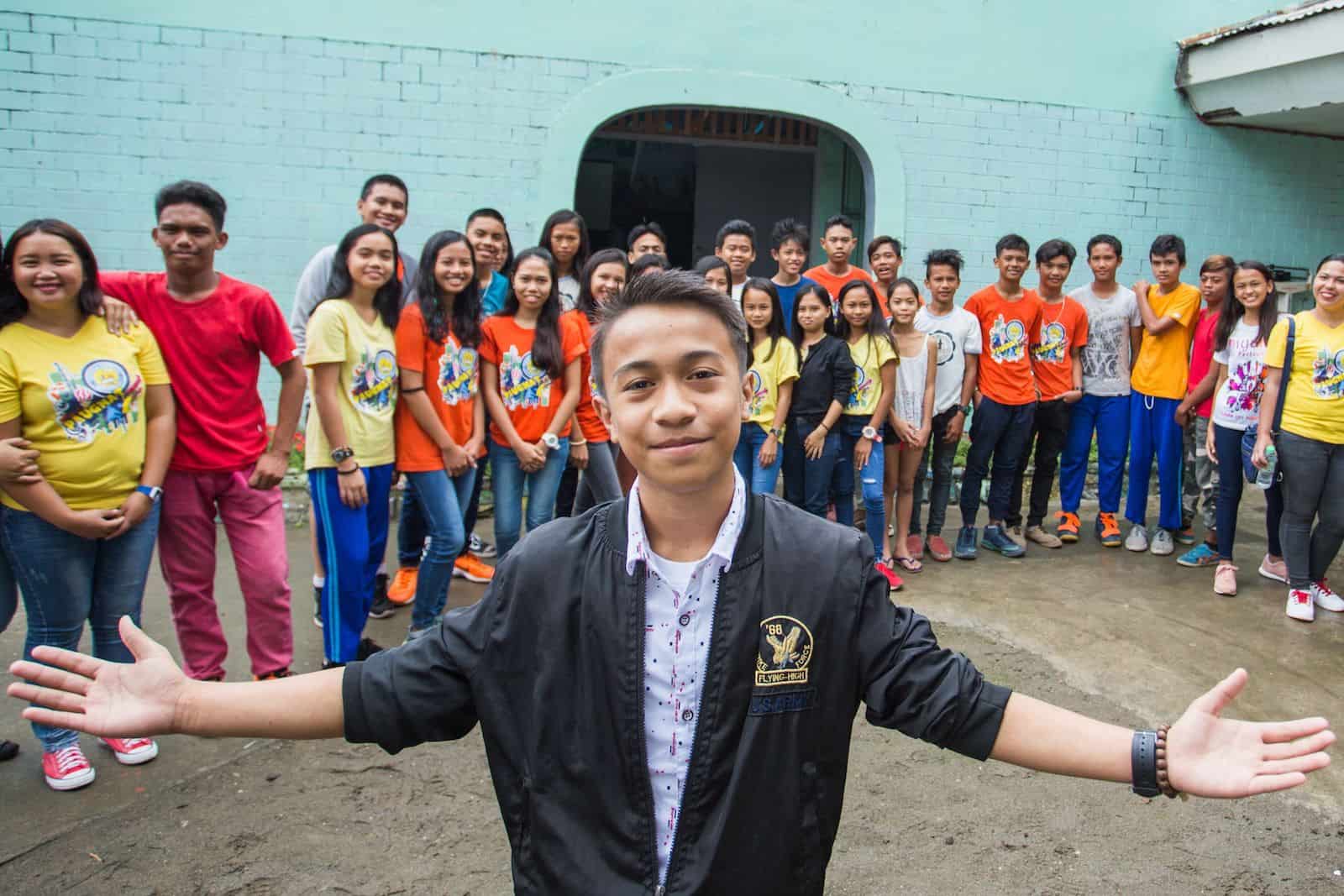 A teen boy, the "Most Excellent Child in the Philippines," wearing a black jacket and white shirt holds his arms out to the side, smiling, standing in front of a large group of children in front of a church.