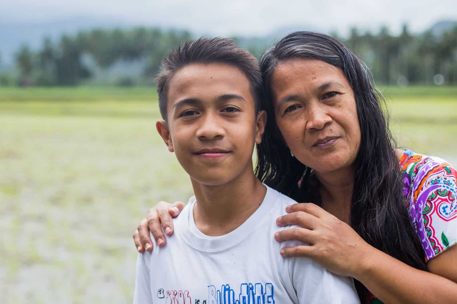 A woman has her arms around a teen boy in a white shirt, standing in front of a green field. 