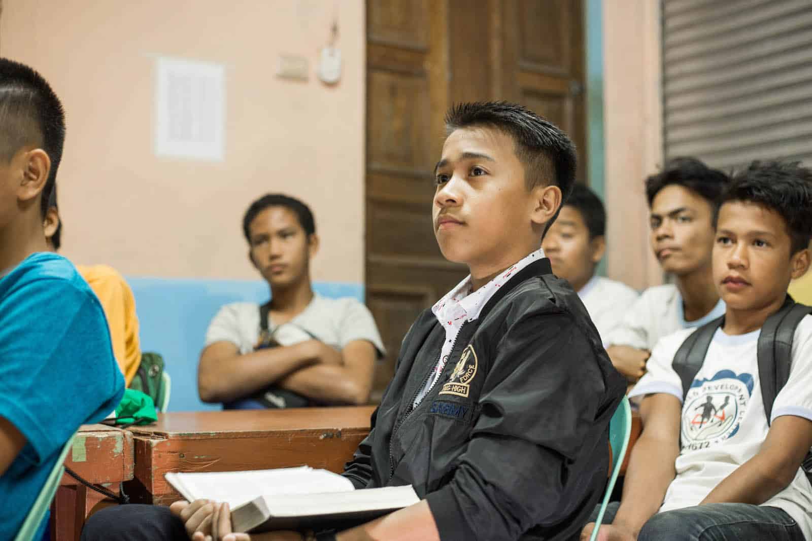 A teen boy sits in a classroom, wearing a black jacket, surrounded by other boys. 