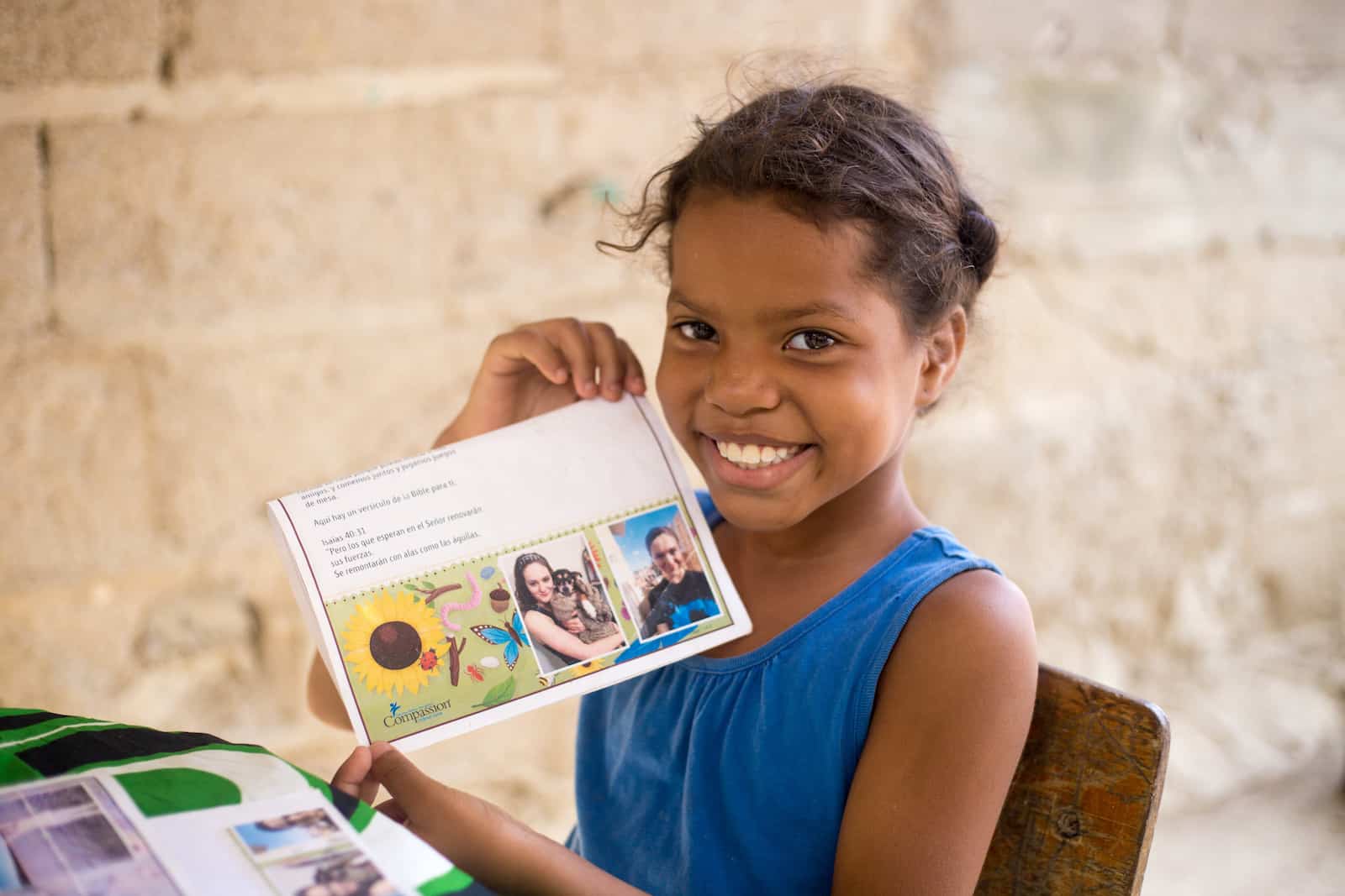 A girl in a blue shirt sits at a table, holding a letter with a picture of people and a sunflower.