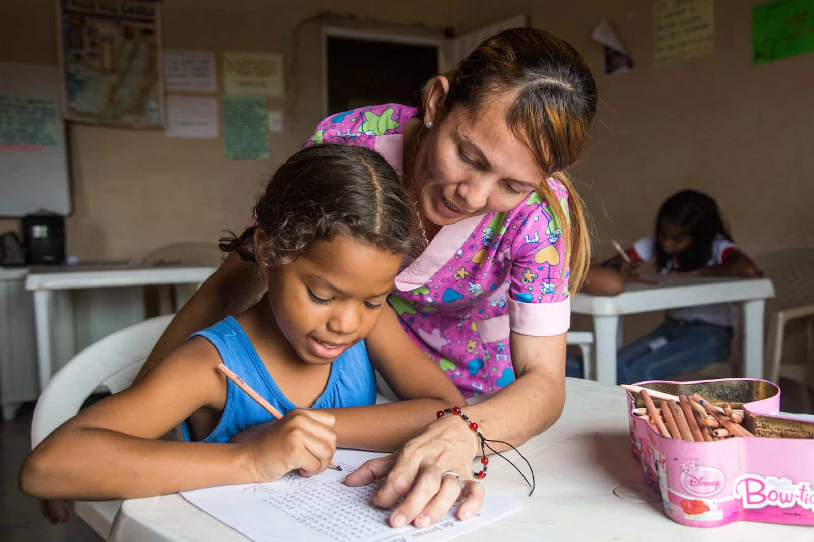 A woman in a pink shirt leans over a girl in a blue shirt writing at a table in a classroom. 