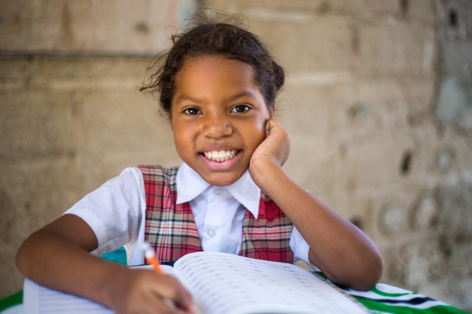 A girl in a red plaid dress over a white collared shirt sits at a table with a pencil in hand, smiling.